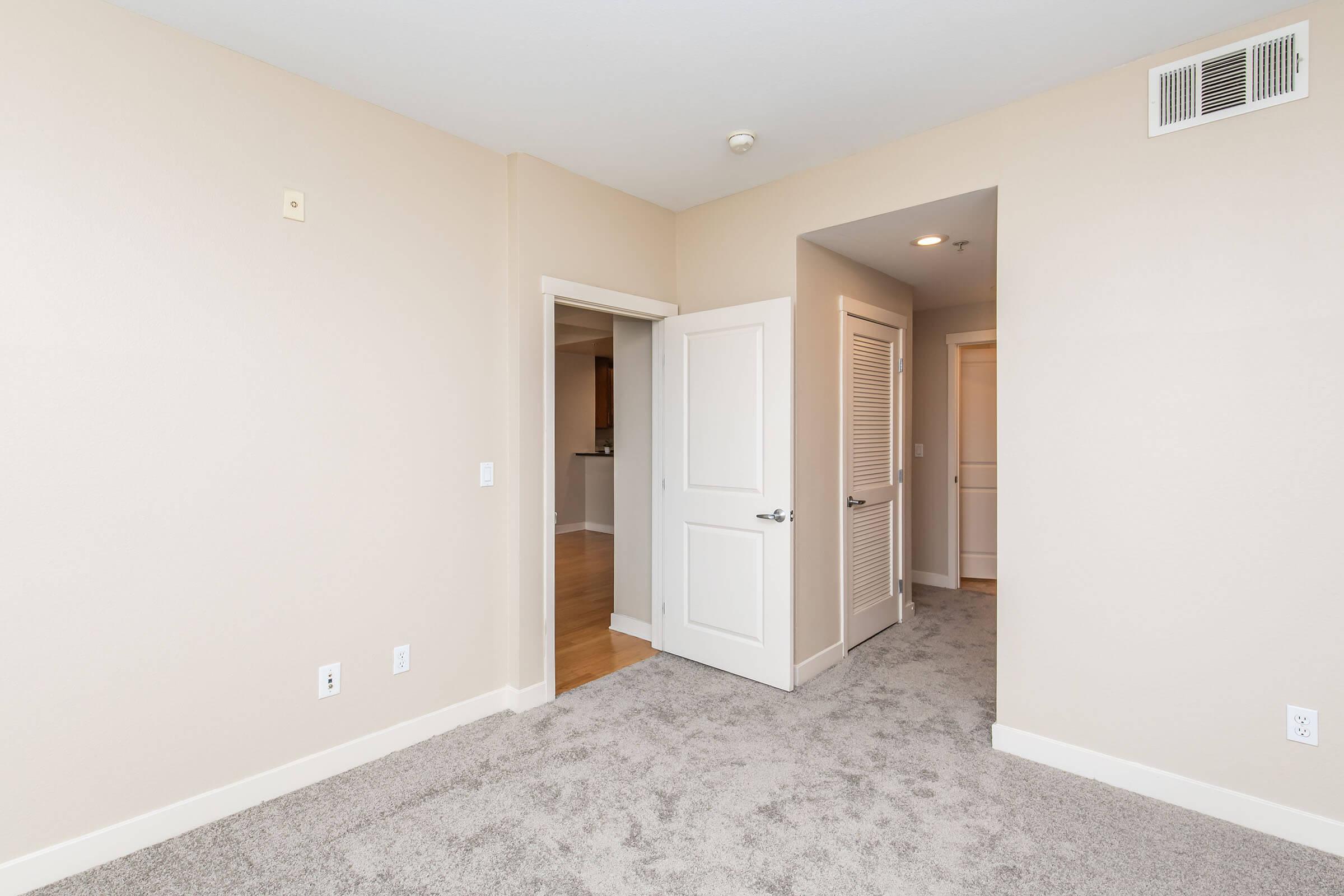 A softly lit interior of a room featuring beige walls and gray carpet. To the left, an open doorway leads to another space, while a second doorway is partially visible on the right. The room has a clean, modern design with no furniture, highlighting its spaciousness.
