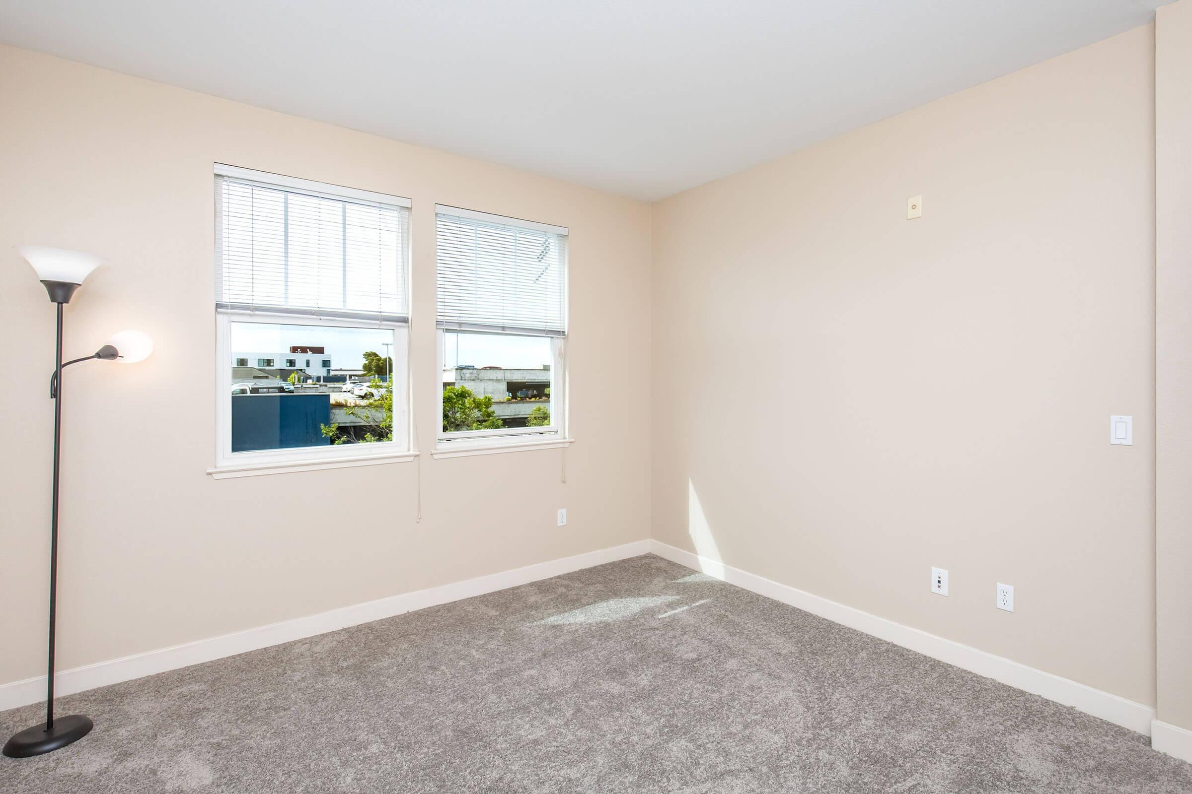 A bright, empty room with light beige walls and a carpeted floor. The room has two windows with white blinds, allowing natural light to come in. A modern floor lamp stands in one corner, adding a touch of elegance. The overall feel is spacious and inviting, ideal for personalizing.