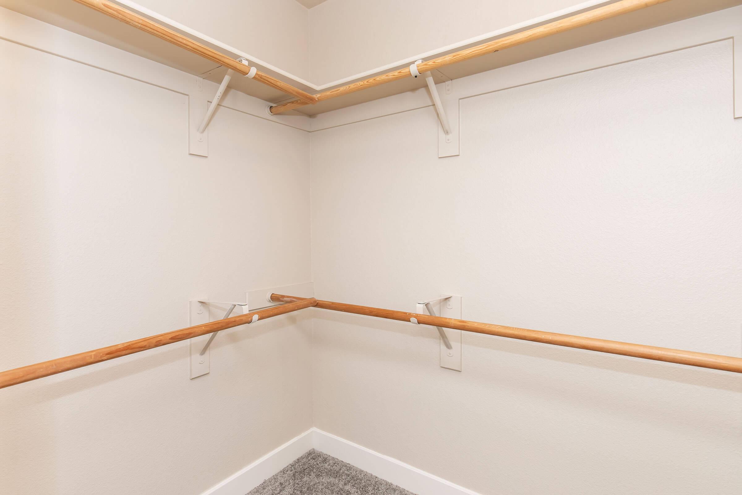 Empty closet with two wooden hanging rods attached to the walls, set against beige walls and a gray carpeted floor. The space is well-lit and minimal, featuring no shelves or additional furniture.