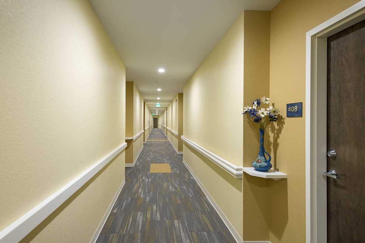 A well-lit hotel corridor with light yellow walls and patterned carpet. There is a blue flower vase on a ledge next to a door labeled "408." The hallway extends in both directions, leading to more doors and a distant exit, with fluorescent lighting illuminating the space.