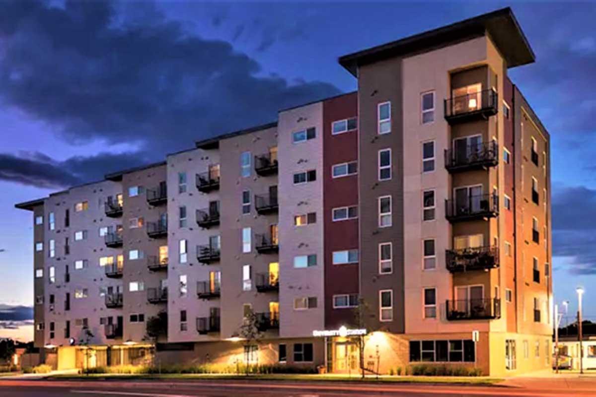 Modern four-story apartment building at dusk, featuring multiple balconies, large windows, and a contemporary facade. The exterior is a mix of neutral colors, with a partially cloudy sky above. Outdoor lighting highlights the entrance and surrounding landscaping.