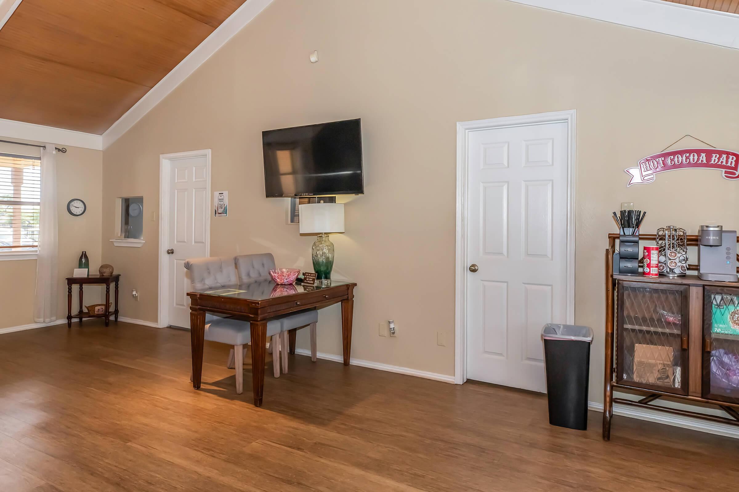A cozy interior space featuring a wooden table with chairs, a television mounted on the wall, and a decorative cocoa bar. The walls are a warm beige color, with a vaulted ceiling made of light wood. A trash bin, decorative items, and a window with blinds add to the welcoming atmosphere.