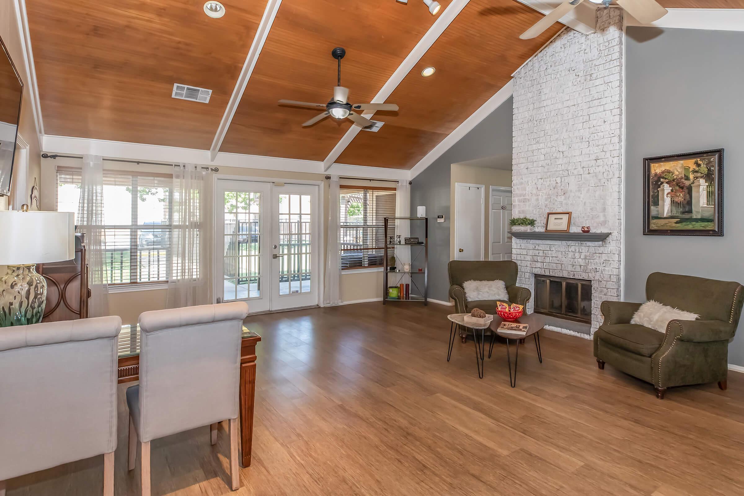 Spacious living room featuring a high vaulted ceiling with wood paneling, two comfortable green armchairs, a coffee table with snacks, and a cozy brick fireplace. Large windows provide natural light, and there's a small desk and a bookshelf in the background, creating an inviting atmosphere.