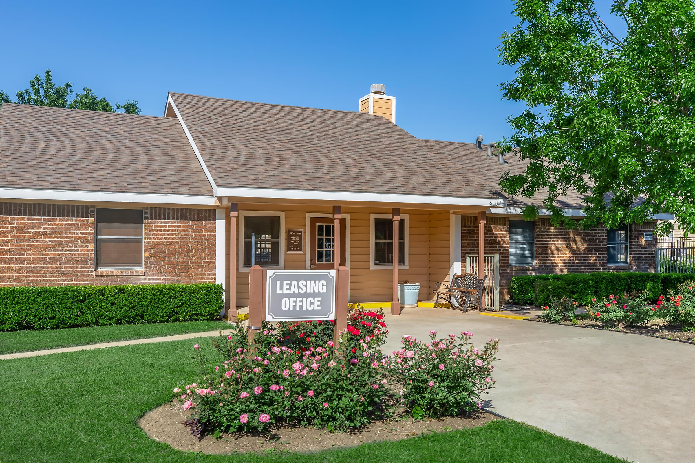 A charming leasing office with a brick exterior and light orange siding. The entrance features a sign reading "Leasing Office" surrounded by well-maintained greenery and blooming rose bushes. The pathway leading to the entrance is paved, and there are comfortable chairs on the porch, inviting visitors to relax.