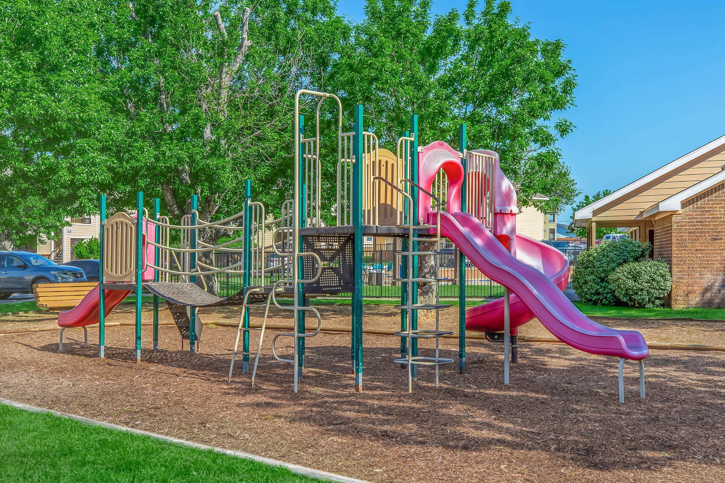 A playground featuring colorful slides, climbing structures, and various play equipment, set on a mulch surface surrounded by green grass and trees. In the background, there are residential buildings and parked cars. The scene is bright and sunny, ideal for outdoor play.