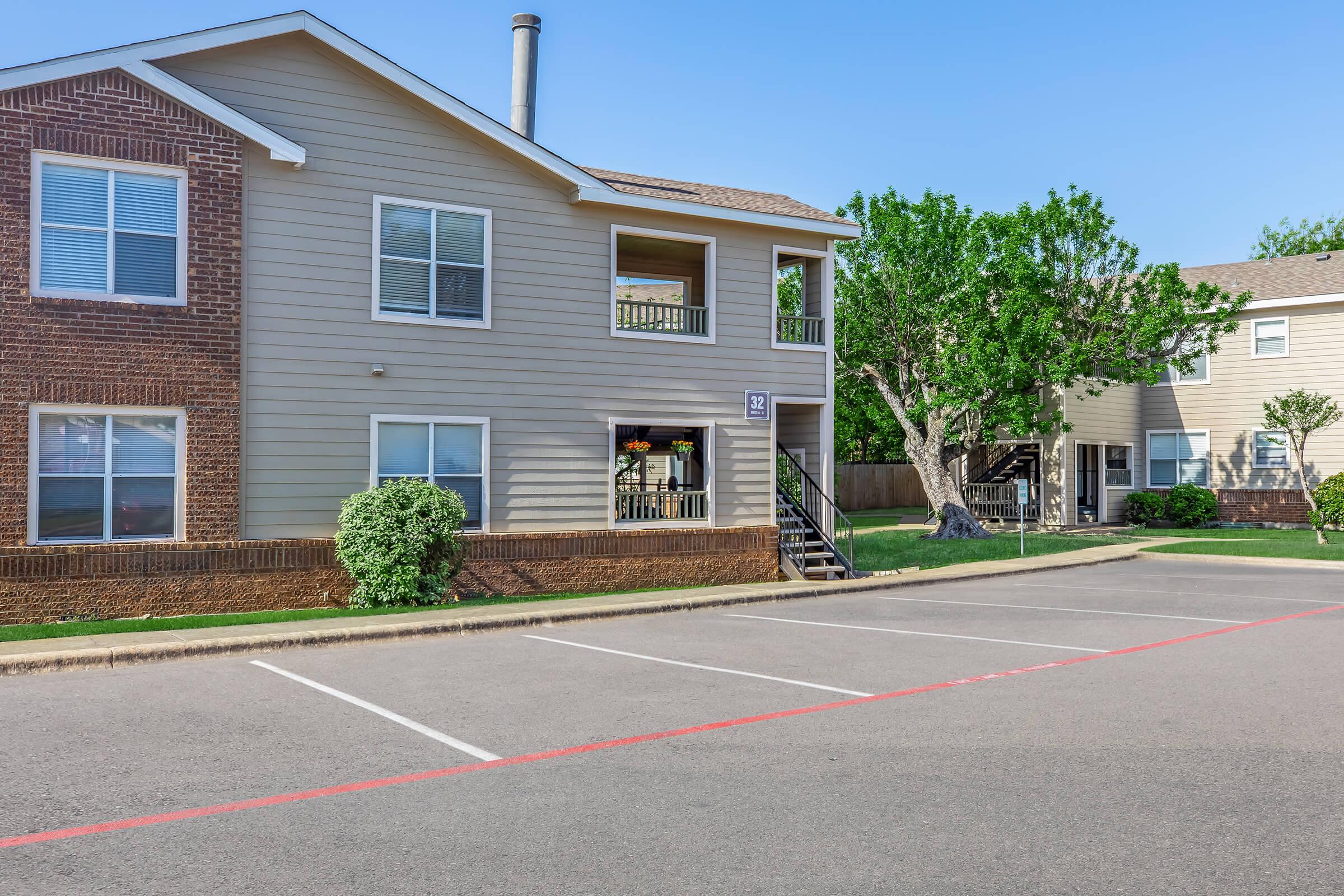 A two-story apartment building with tan siding and brick accents, featuring several windows and a balcony. The parking lot in front has empty spaces marked by red lines, while trees and shrubs are visible in the surrounding landscape. Clear blue sky in the background.