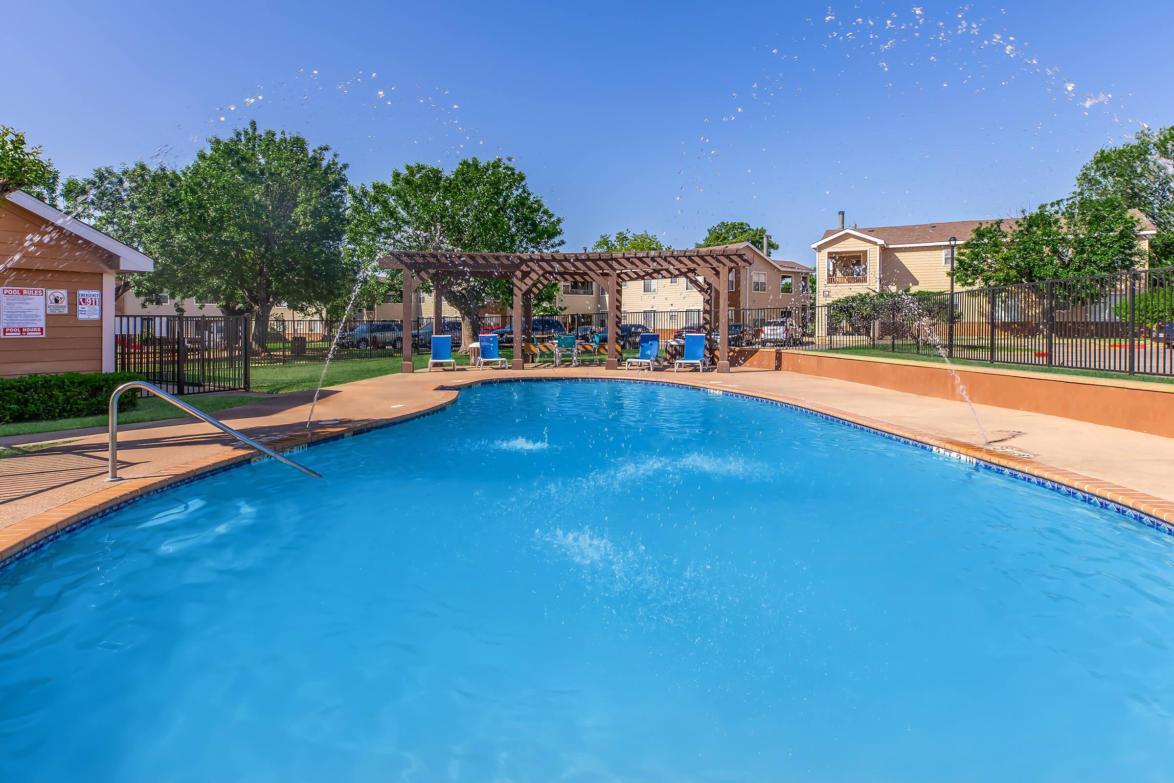 A bright outdoor swimming pool surrounded by greenery, featuring a pergola and lounge chairs. Water features create splashes in the clear blue water. Nearby, there are residential buildings and a well-maintained lawn, showcasing a sunny day and a relaxing atmosphere.