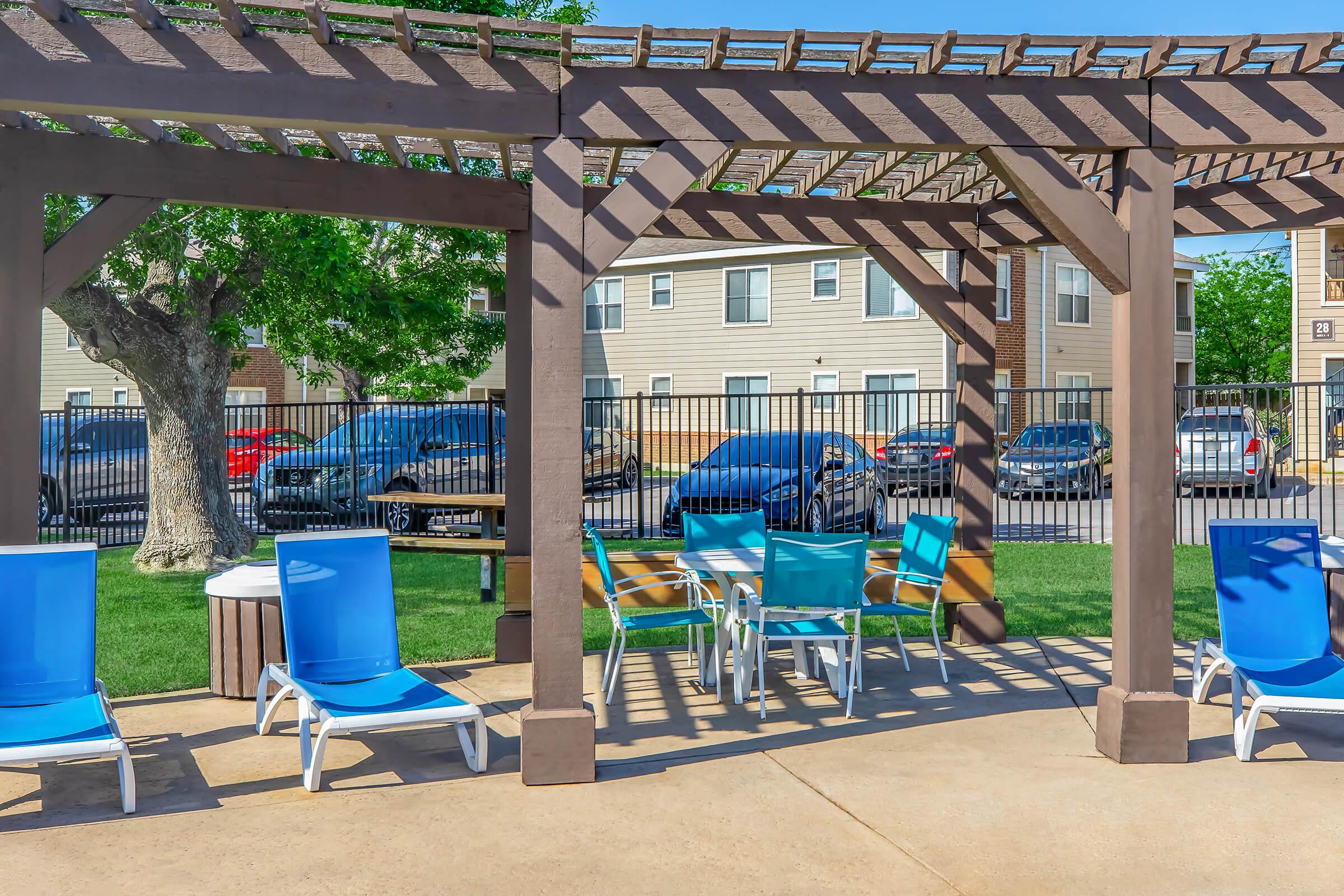 A shaded area by a pool featuring wooden pergolas, blue lounge chairs, and a table with four chairs. Surrounding the area are green lawns and a fence, with parked cars visible in the background and residential buildings in the distance.
