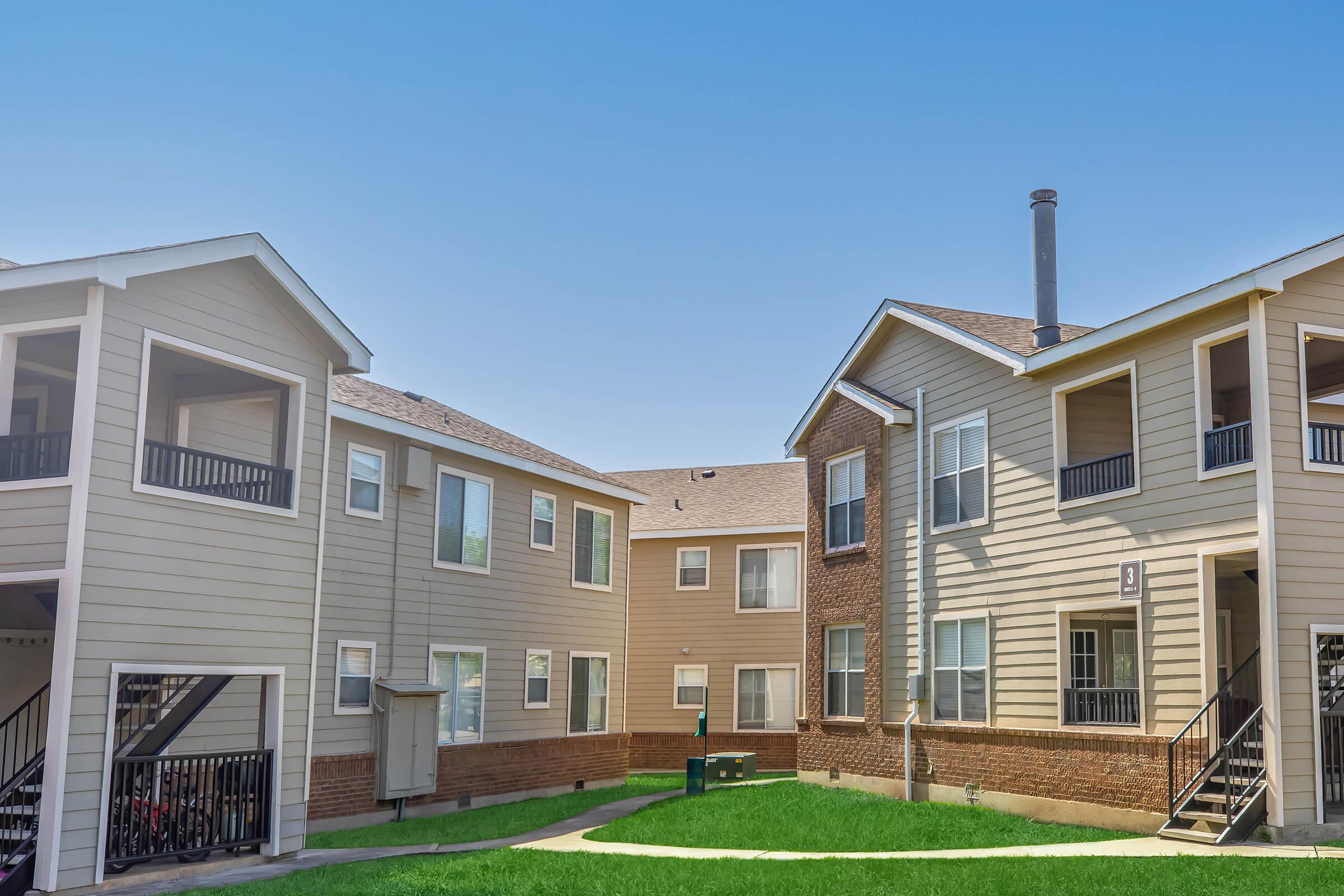 Two multi-story residential buildings with light-colored siding and brick accents, surrounding a grassy courtyard. The buildings feature balconies and large windows. A walkway leads through the green lawn, and there are staircases visible on the exteriors. The sky is clear and blue.