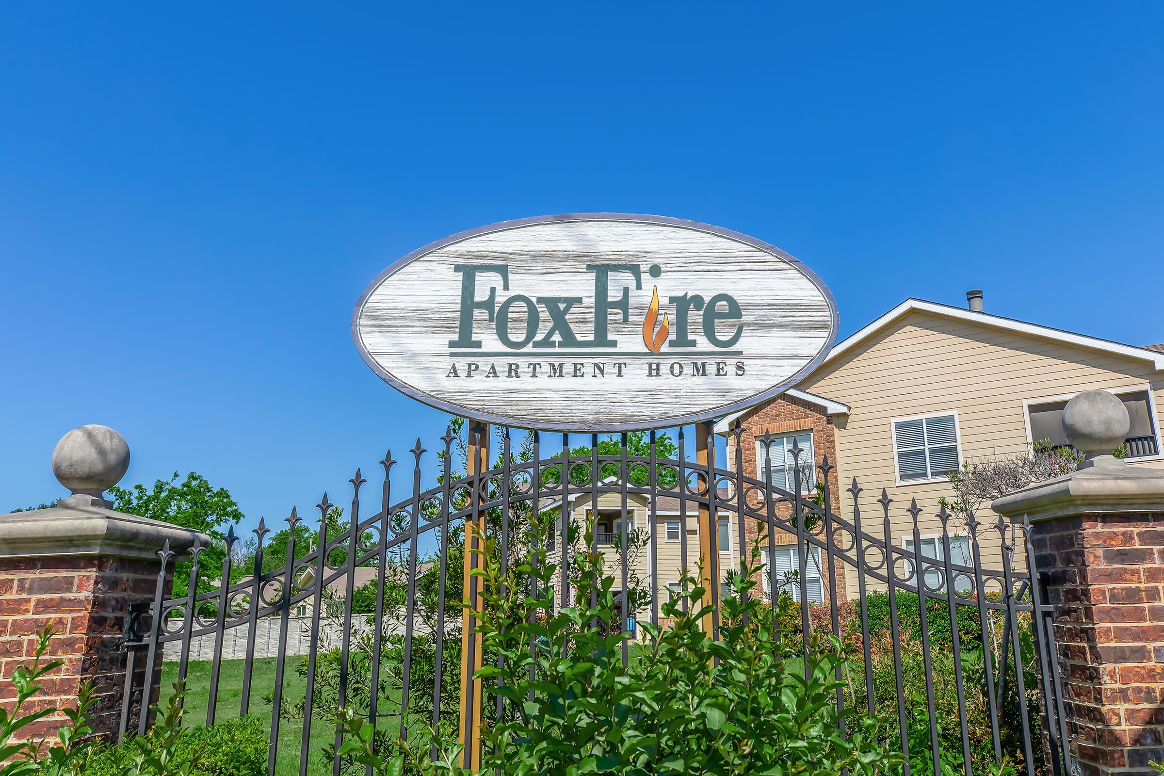 Sign for FoxFire Apartment Homes, featuring a wooden design with the name "FoxFire" prominently displayed. The sign is supported by an ornate gate, with a clear blue sky in the background and a residential building visible behind the gate.