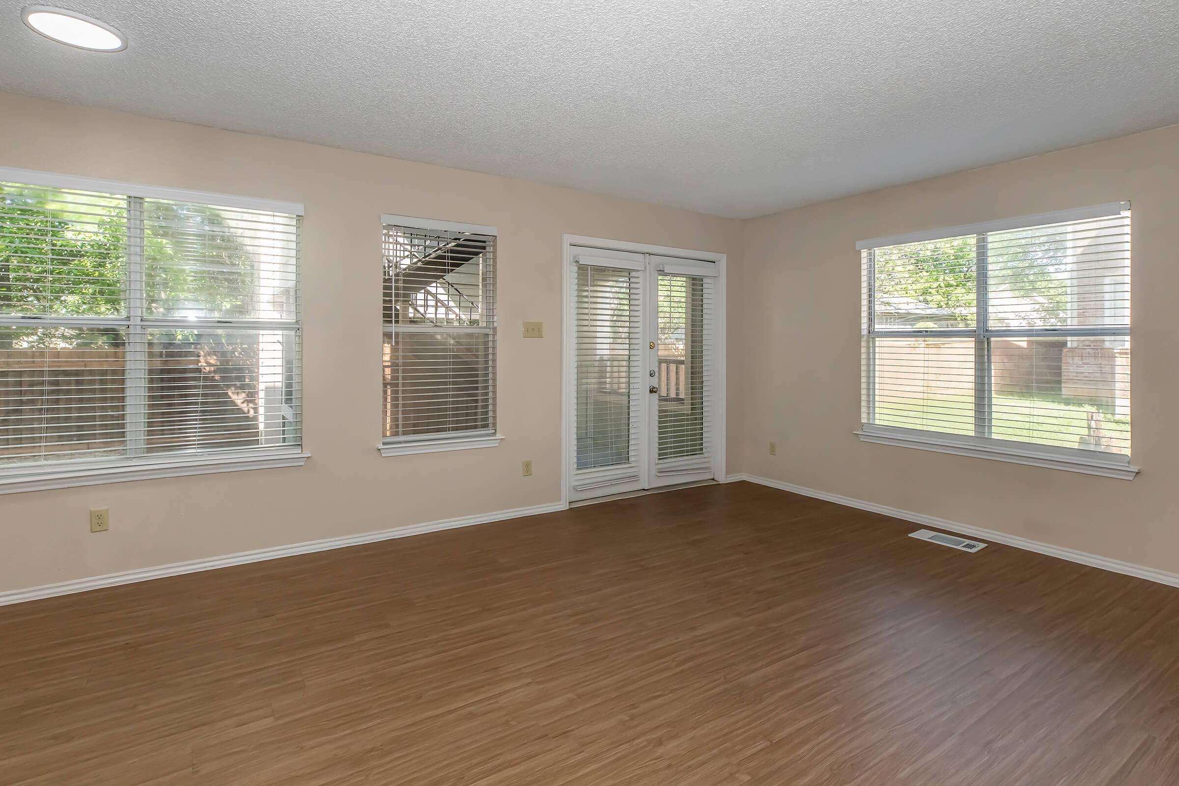 Bright and spacious interior with light-colored walls, wooden flooring, and large windows allowing natural light. The room features sliding glass doors leading to an outdoor area, partially visible, and includes window blinds.