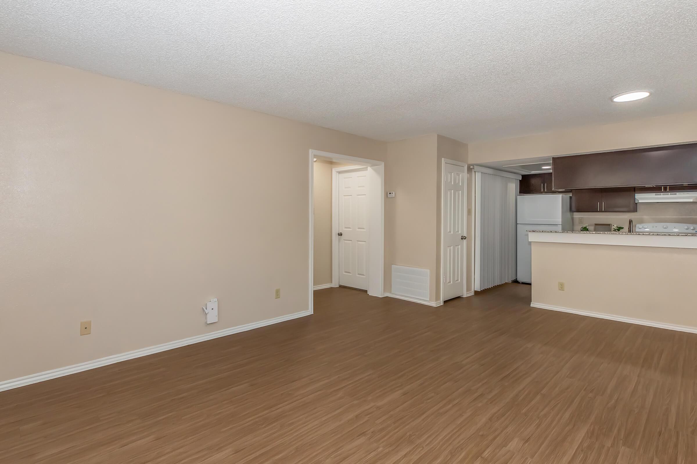 Spacious, empty living area with light-colored walls and a textured ceiling. A doorway leads to another room, and the kitchen is partially visible with dark cabinetry and appliances. The floor features light wood laminate, creating a bright and open atmosphere.