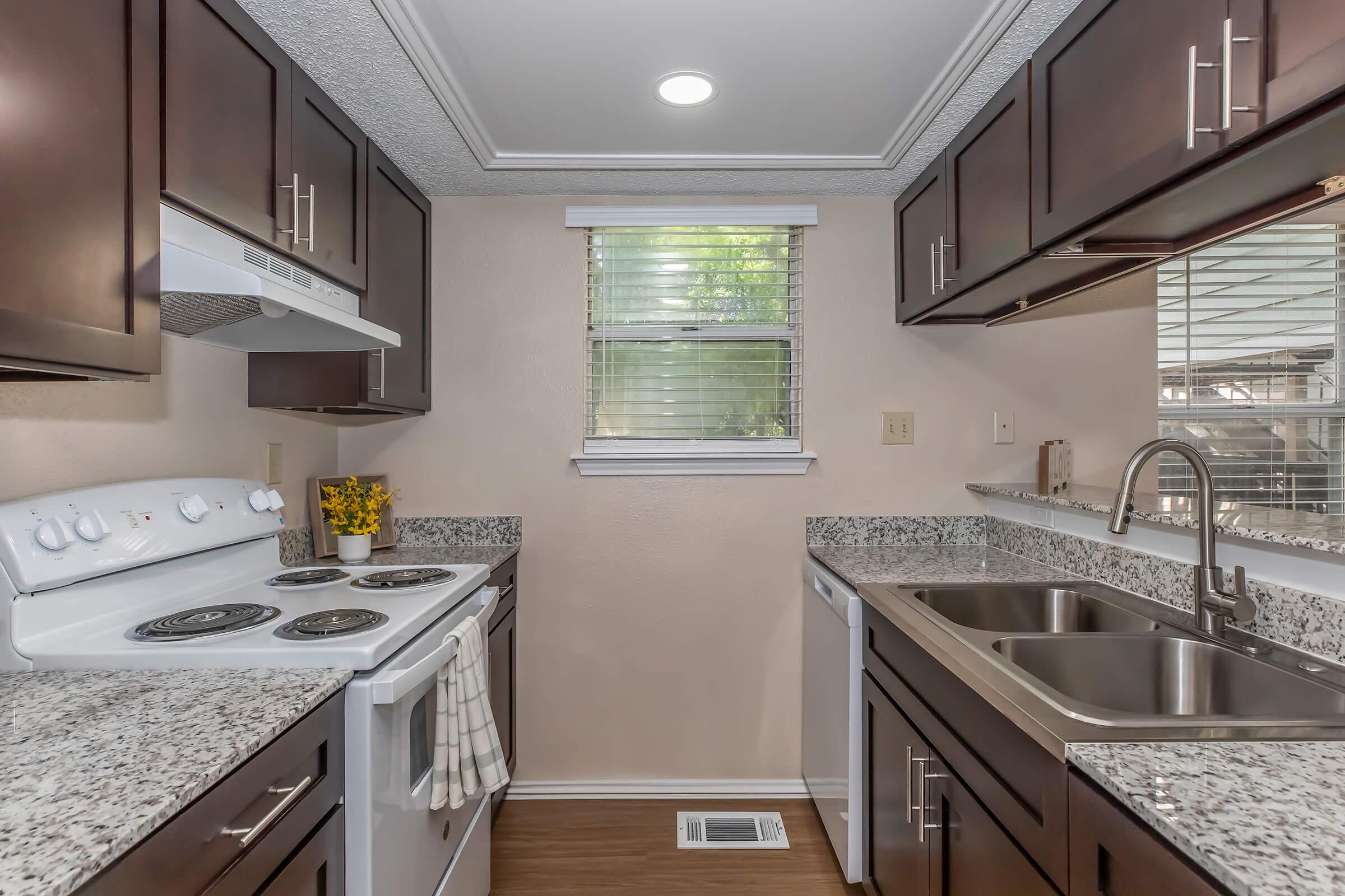 Modern kitchen featuring dark wood cabinetry, granite countertops, a white stove, and a double sink. Natural light filters in through a window with blinds. The floor is a light wood laminate, and there is a small vase of yellow flowers on the counter. A dishwasher is also visible.
