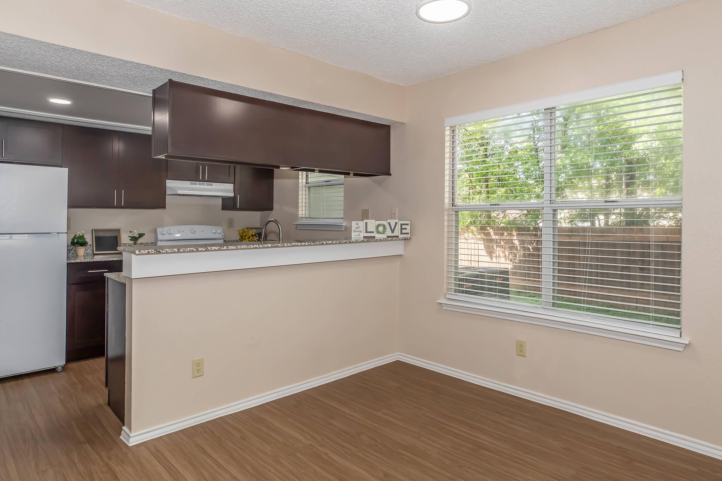 A bright and open kitchen area featuring dark wooden cabinets, a white countertop, and a window with blinds. Light-colored walls enhance the spacious feel, and the floor is made of light wood. A decorative sign spelling "LOVE" is displayed on the countertop.