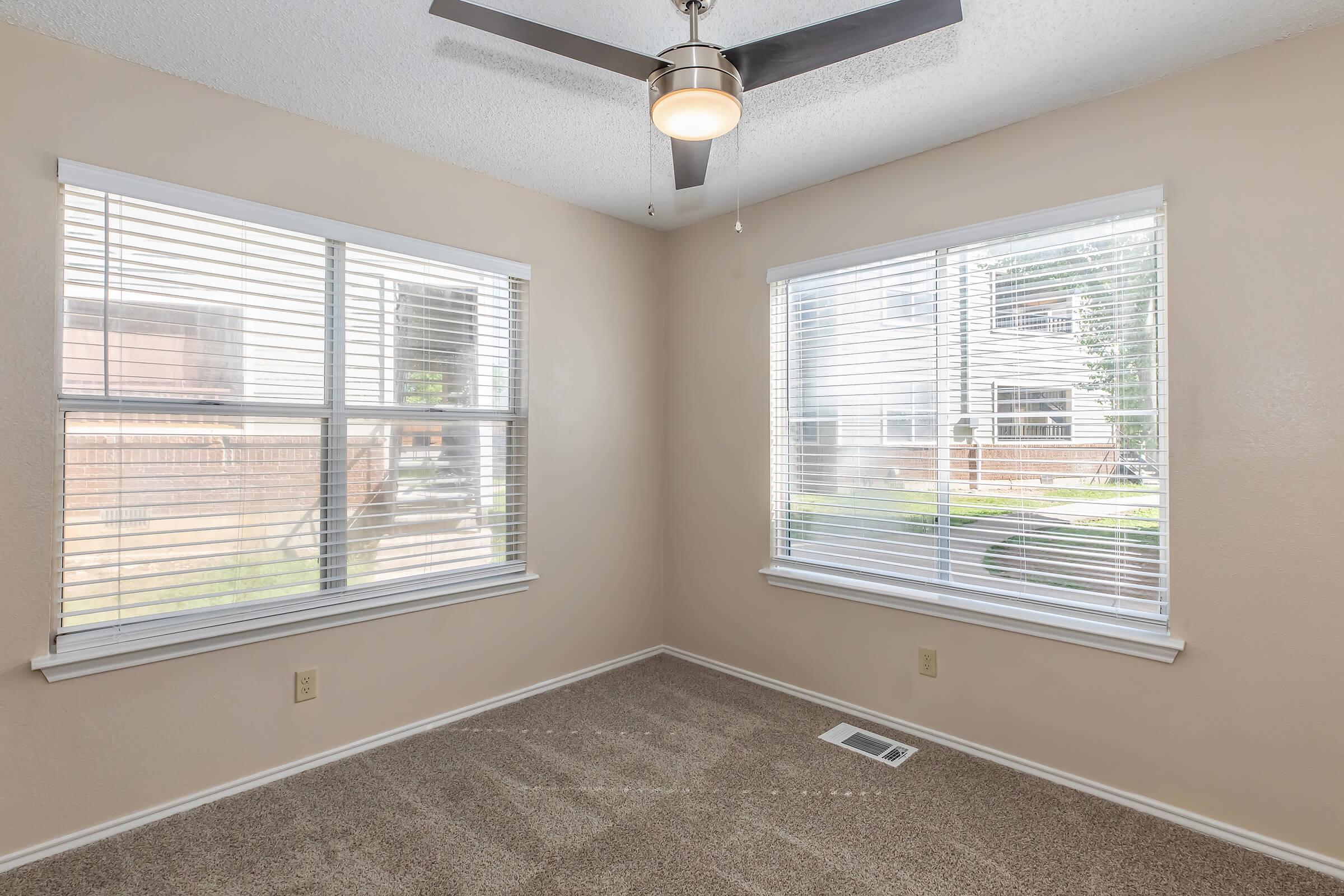 Bright corner room featuring two large windows with blinds, a ceiling fan, and freshly carpeted flooring. The walls are a neutral color, enhancing the spacious feel. Outside the windows, a glimpse of greenery is visible, suggesting a pleasant outdoor area.