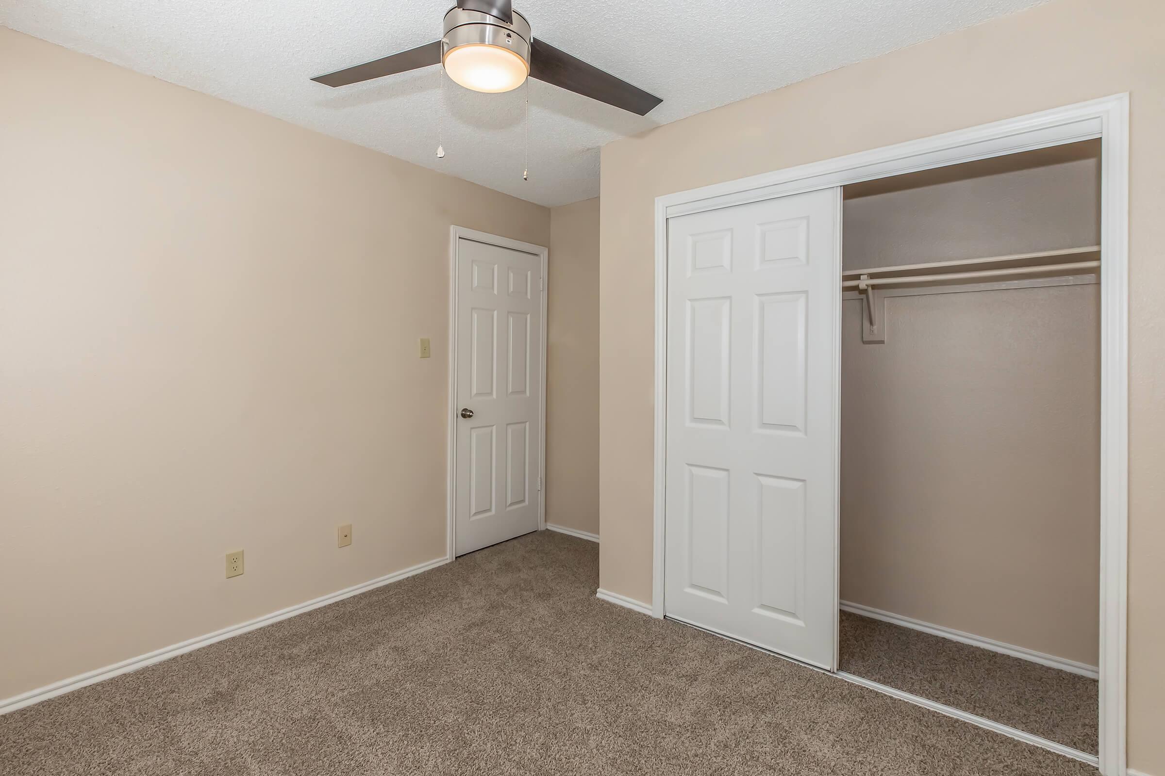 A beige room with a ceiling fan, featuring a closet with double doors and plush carpet flooring. The walls are painted a light color, and there is a single door leading to another space. The lighting is bright and inviting, creating a simple and clean aesthetic.