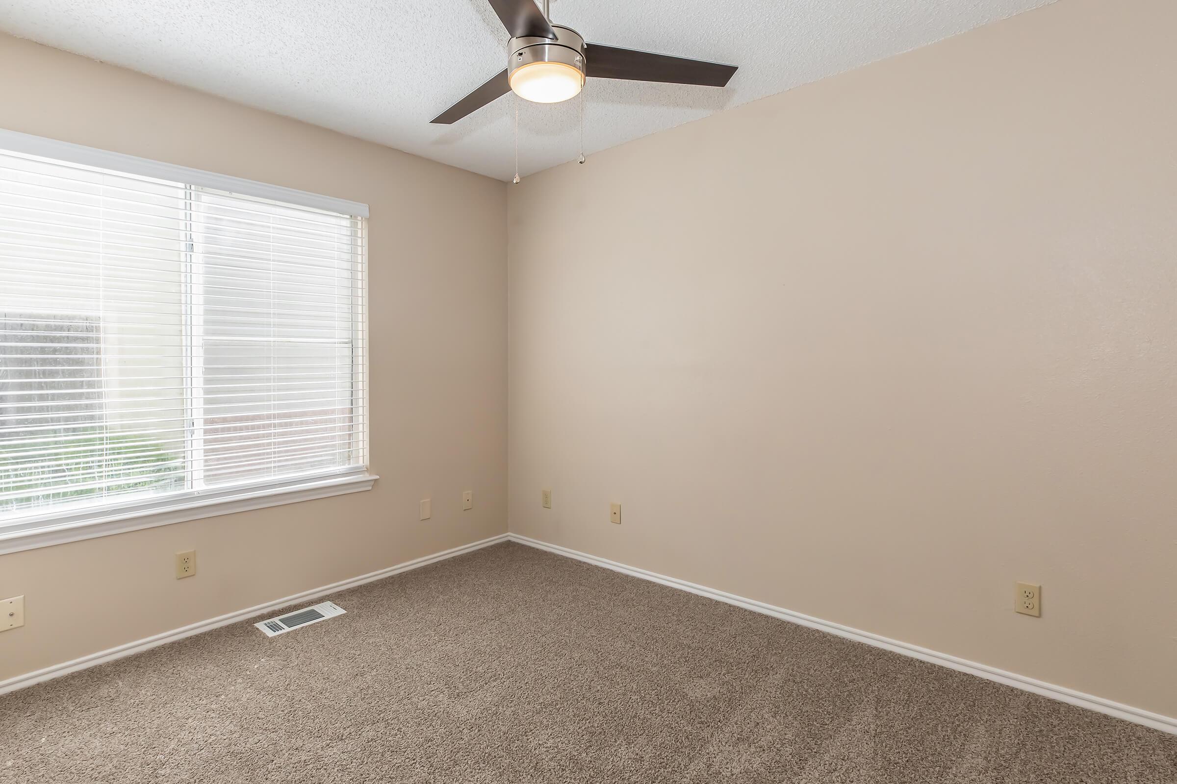 Empty room featuring light beige walls and carpet. A ceiling fan is mounted in the corner, and a window with white blinds provides natural light. The floor has a vent, and there are no furnishings or decorations, creating a minimalistic space.