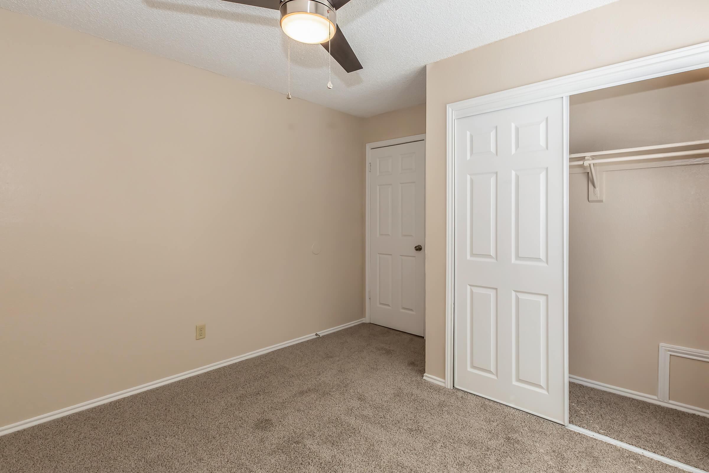 Empty room with light beige walls and carpeted flooring. A ceiling fan is mounted on the ceiling, and there is a closed door leading to a closet on one side. The room has minimal decor, creating a simple and clean aesthetic.