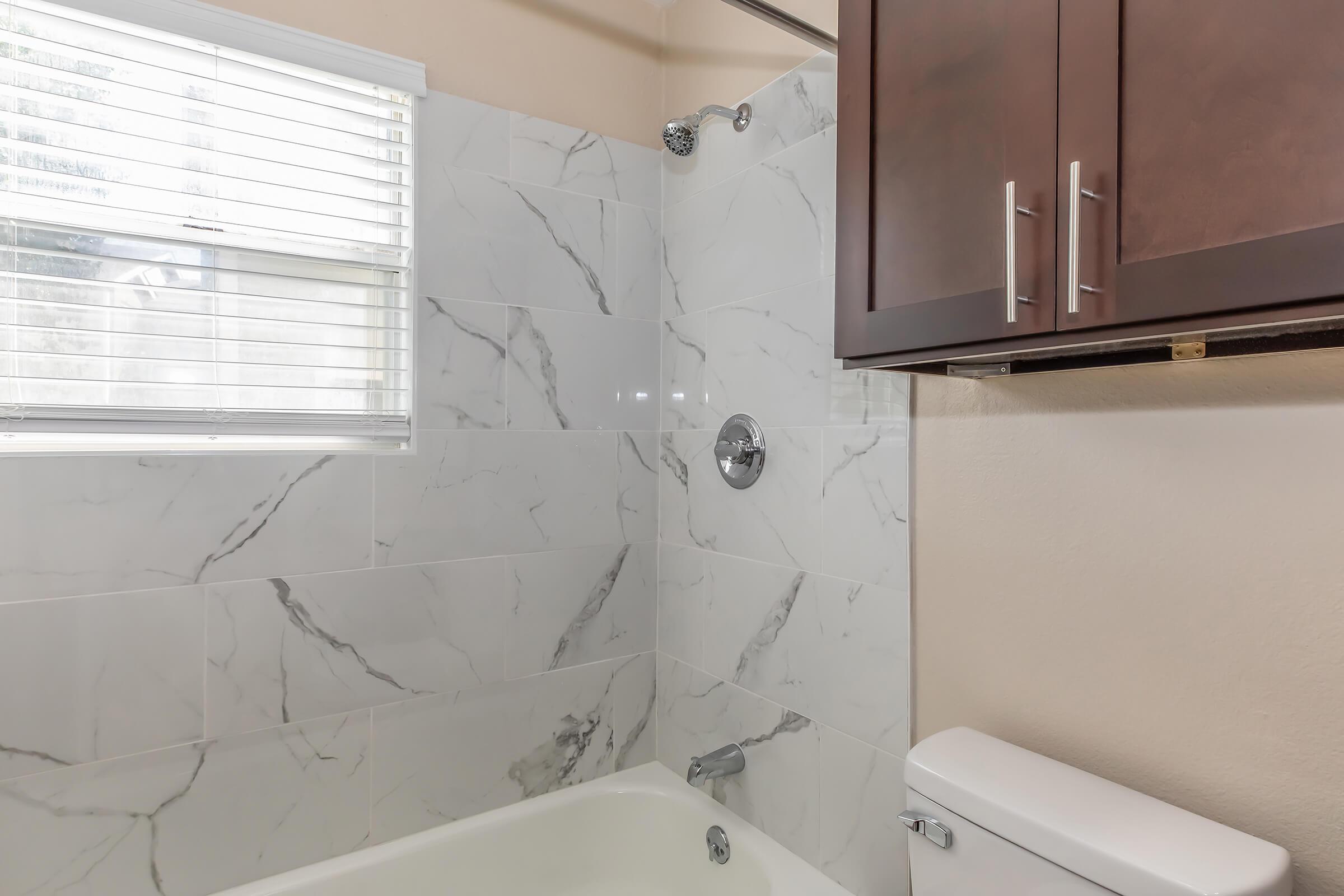 A modern bathroom featuring a shower with a marble-patterned tile wall, a white bathtub, and a toilet. Natural light comes through a window with white blinds. Dark brown cabinetry is visible above the toilet, adding a touch of contrast to the light-colored walls and fixtures.