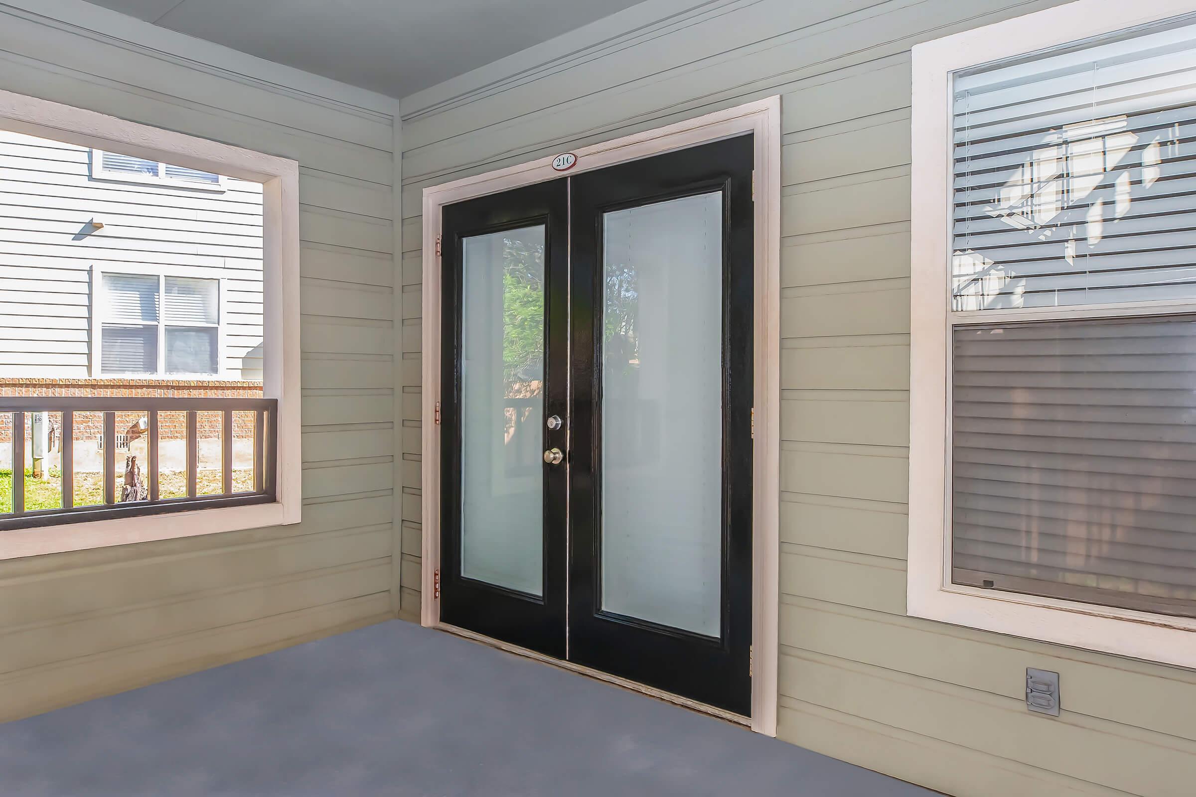 A view of a porch with light green walls and grey flooring. There are large black double doors at the center, flanked by windows with white trim. Natural light filters in from the side, highlighting the simple, modern design of the entrance area.