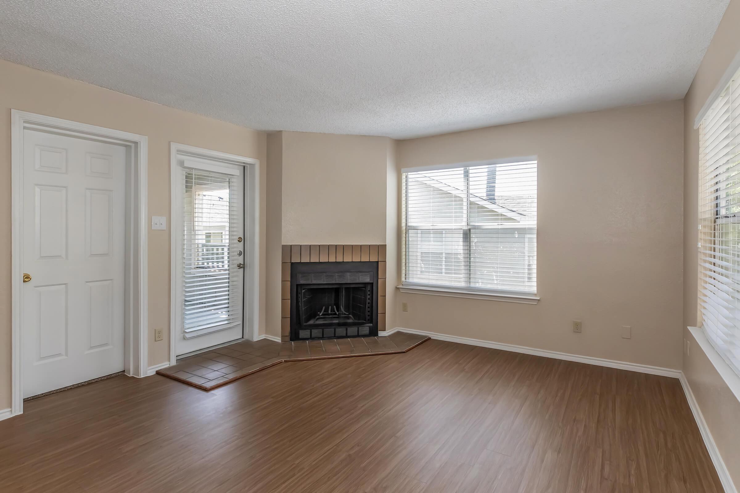 A spacious living room with light beige walls and a textured ceiling. It features a fireplace against one wall, large windows with white blinds allowing natural light, and a door leading outside. The flooring is light wood laminate, creating a warm and inviting atmosphere.