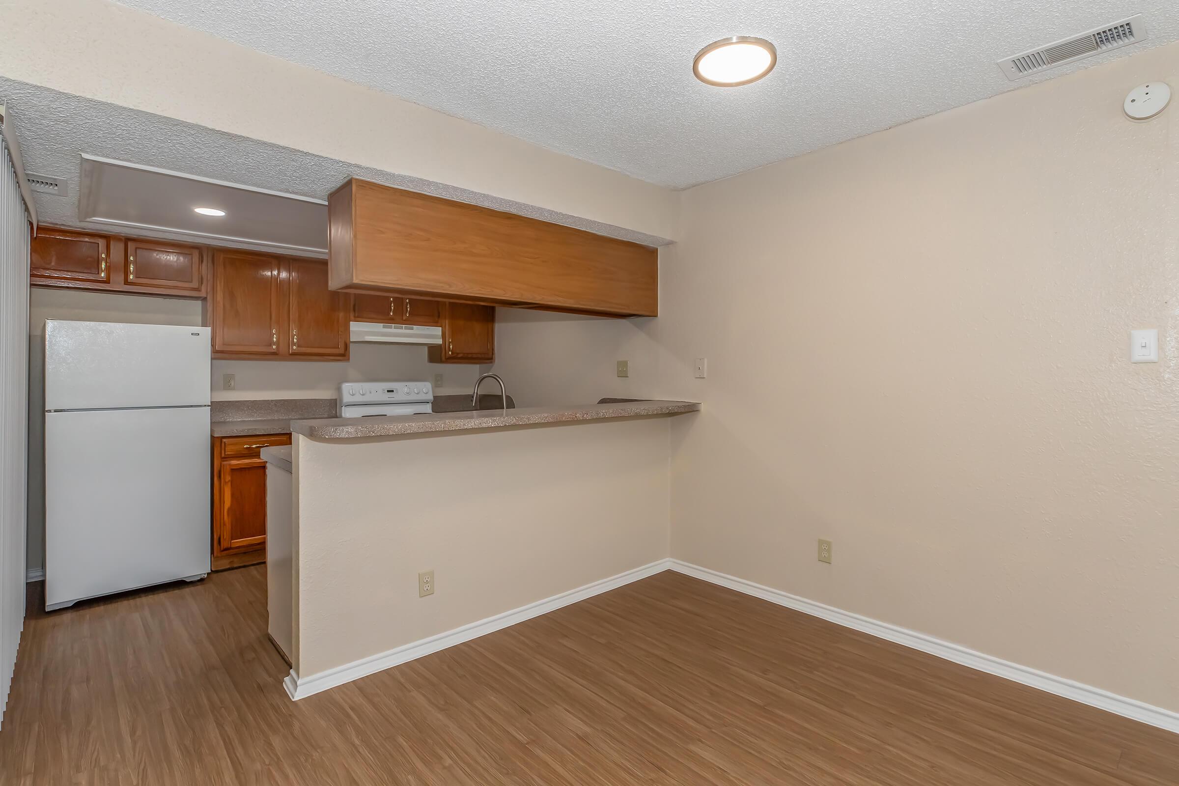 A spacious, light-colored kitchen and living area with a partial wall. The kitchen features wooden cabinetry, a white refrigerator, and a stove. The living area has a wood-like floor and a ceiling light, creating a bright and open atmosphere.