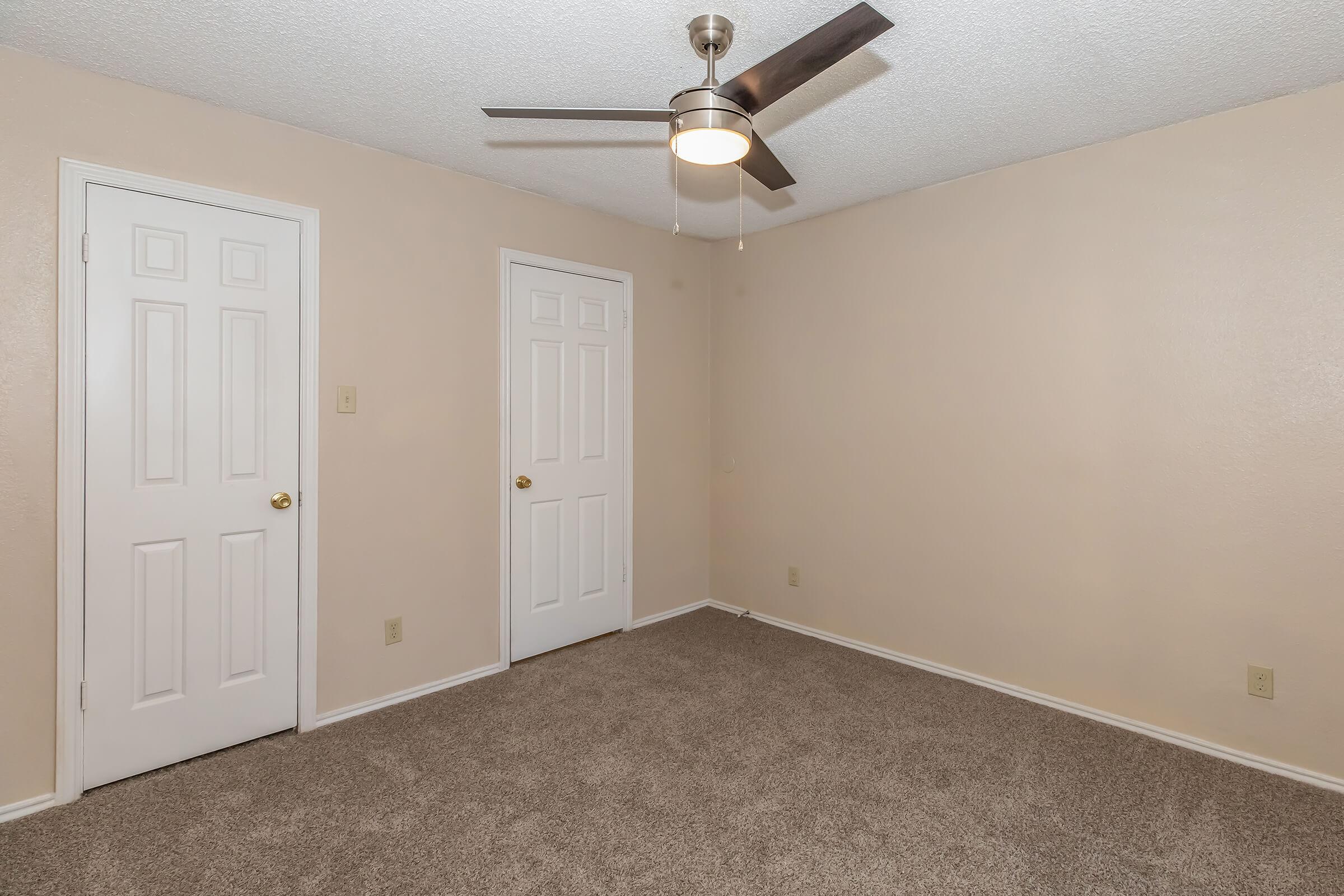 A clean, empty bedroom featuring light beige walls and plush carpet. There’s a ceiling fan with modern design, two closed white doors, and no furniture, creating a spacious and airy atmosphere. Natural light fills the room from an unseen window.