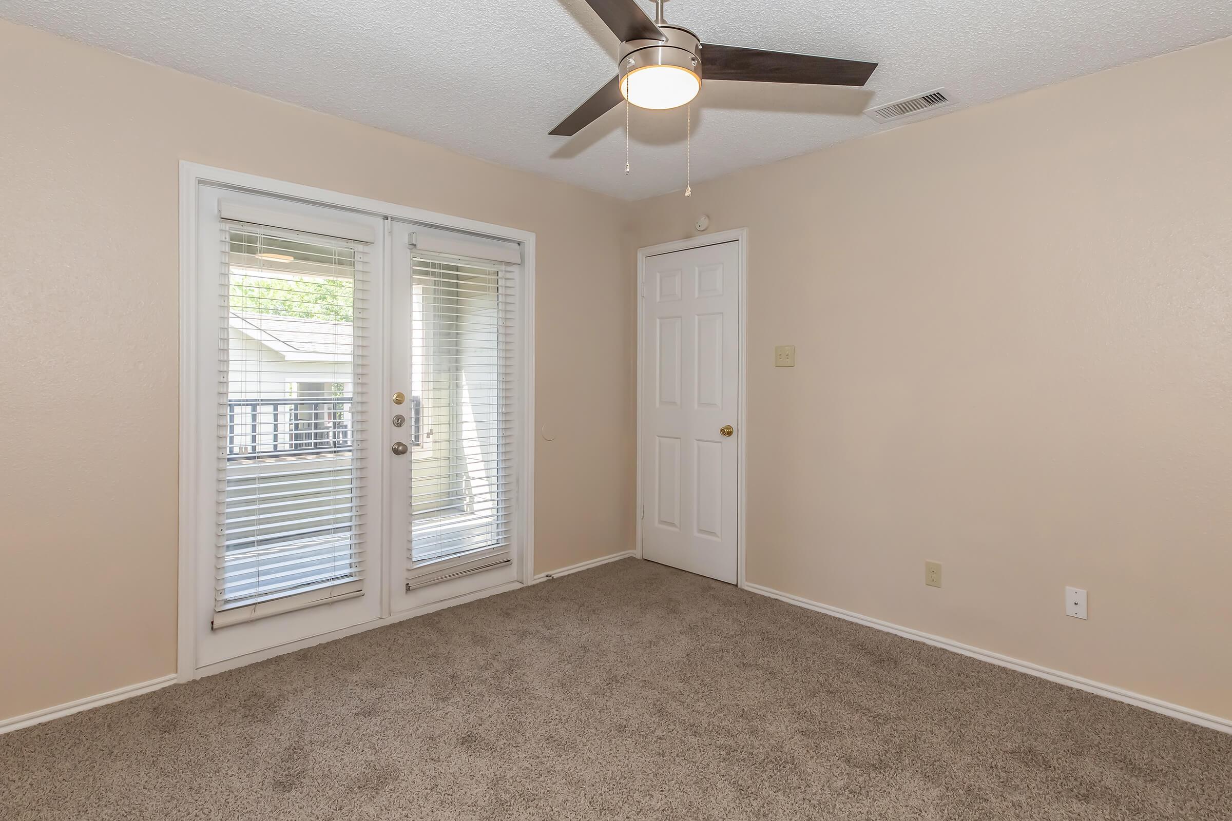 Empty room with beige walls and carpet, featuring a ceiling fan, a set of double doors with blinds leading to a balcony, and a single door on the right. The space is well-lit, showcasing minimal decor and a neutral color palette, creating a cozy atmosphere.
