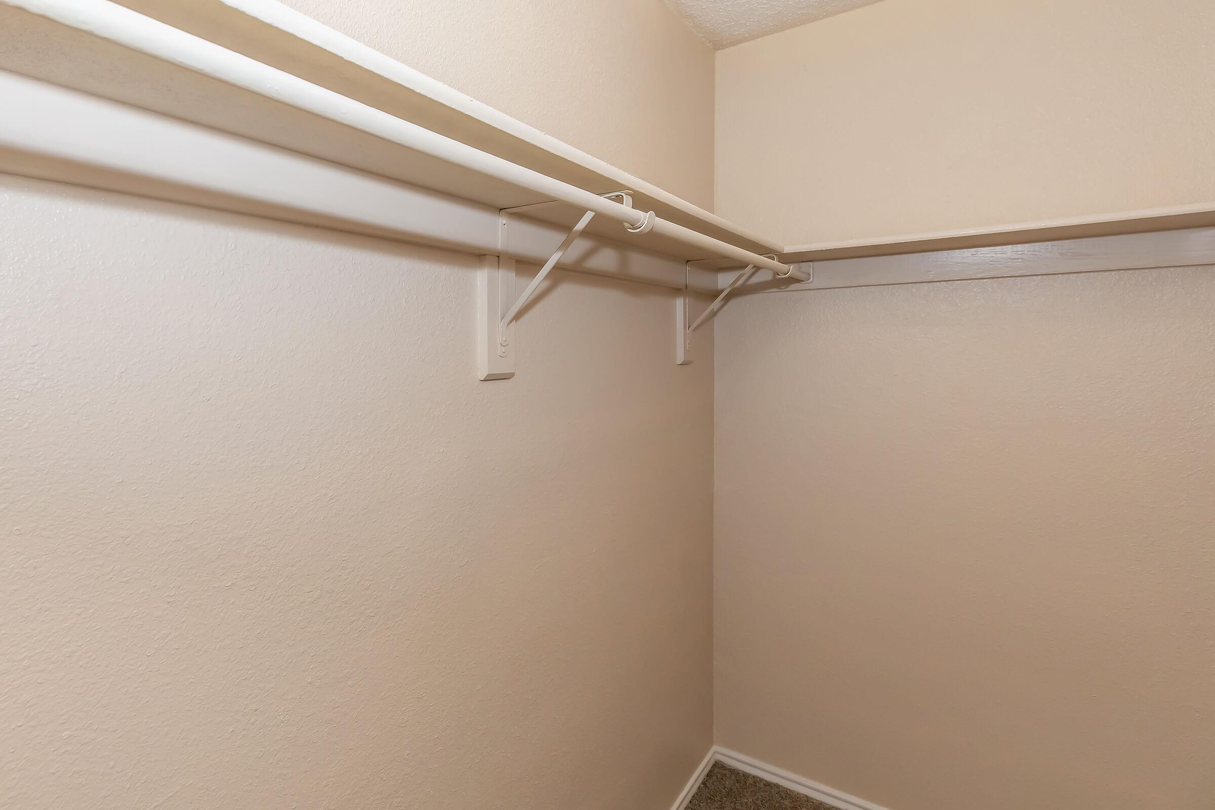 Empty closet corner featuring beige walls and carpeting, with two white hanging rods mounted on brackets. The space appears clean and organized, providing ample room for storage.
