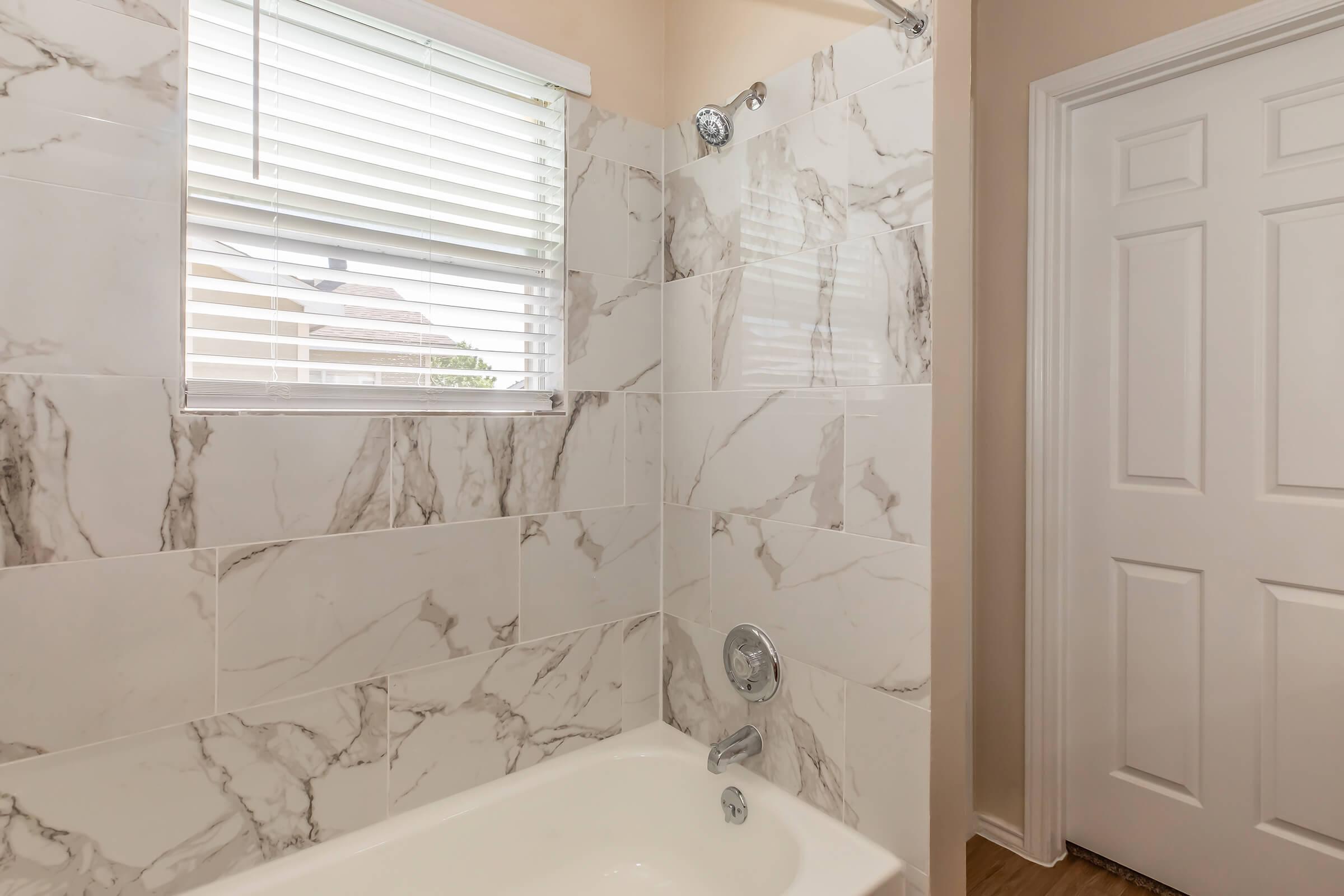 A bathroom featuring a shower/tub combo with a modern design. The walls are tiled with white and gray marble-patterned tiles. A small window with blinds allows natural light in. A white door is visible on the right side, leading to another space. The overall decor is clean and contemporary.