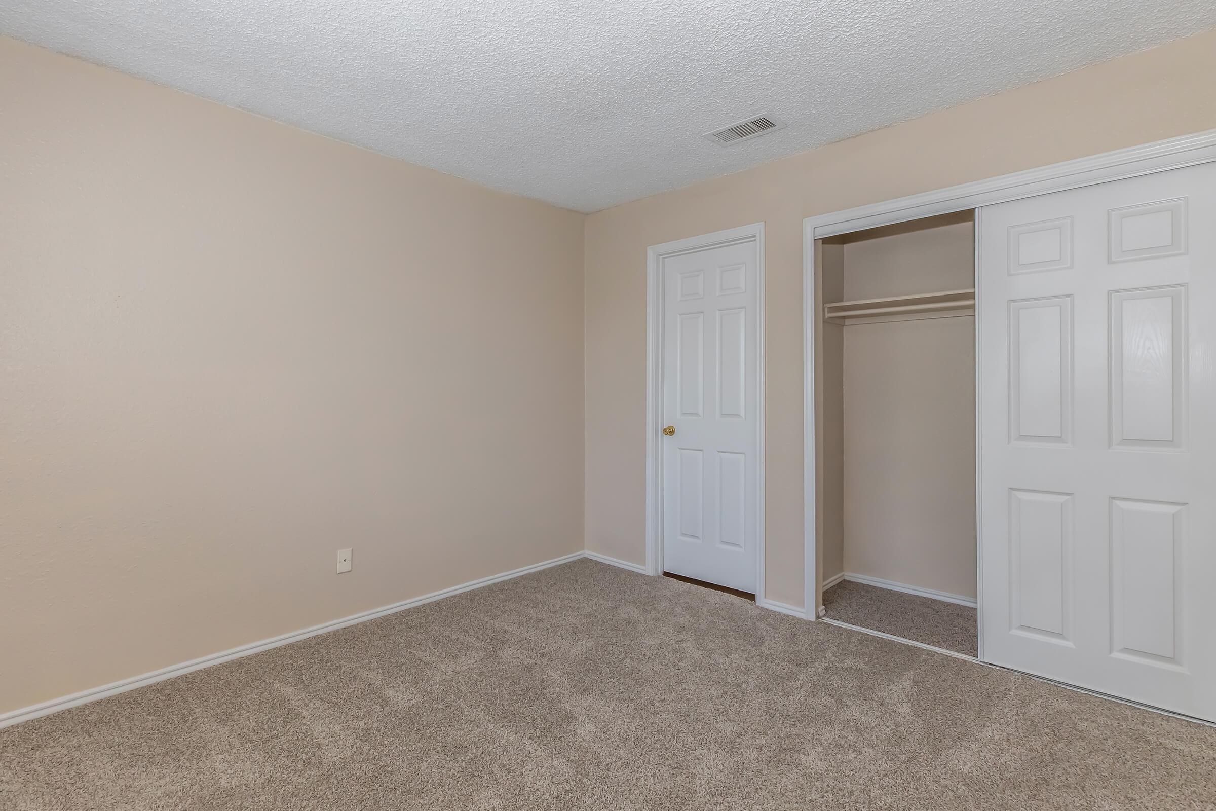 A spacious, empty room with beige walls, soft carpet, and a closet with double doors. There is a single door leading to another area of the home. The room has no furniture, creating a blank canvas for decoration.