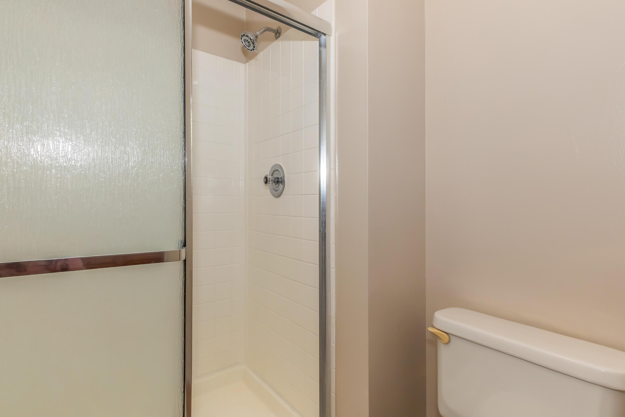 A bathroom featuring a glass shower enclosure with a showerhead and a white tiled interior. To the right, there is a toilet with a simple design, and the walls are painted in neutral tones. The overall appearance is clean and modern.