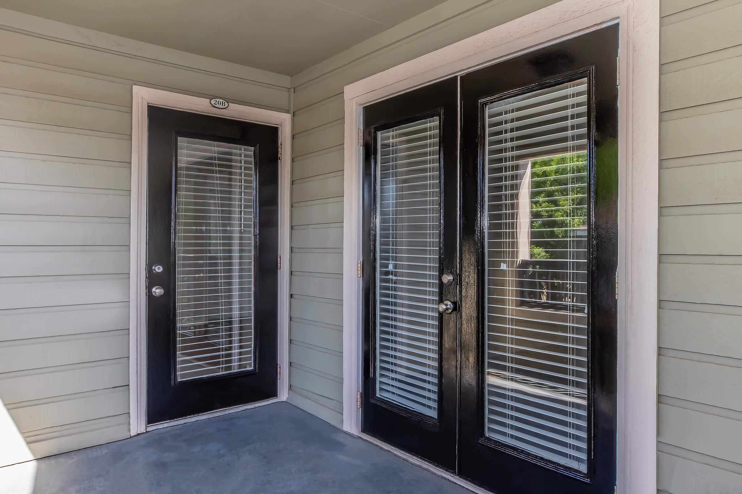 A close-up view of a porch area featuring two black glass doors with vertical blinds, one single door and one double door, set against light green siding. The floor is concrete, and the overall ambiance is inviting and modern.