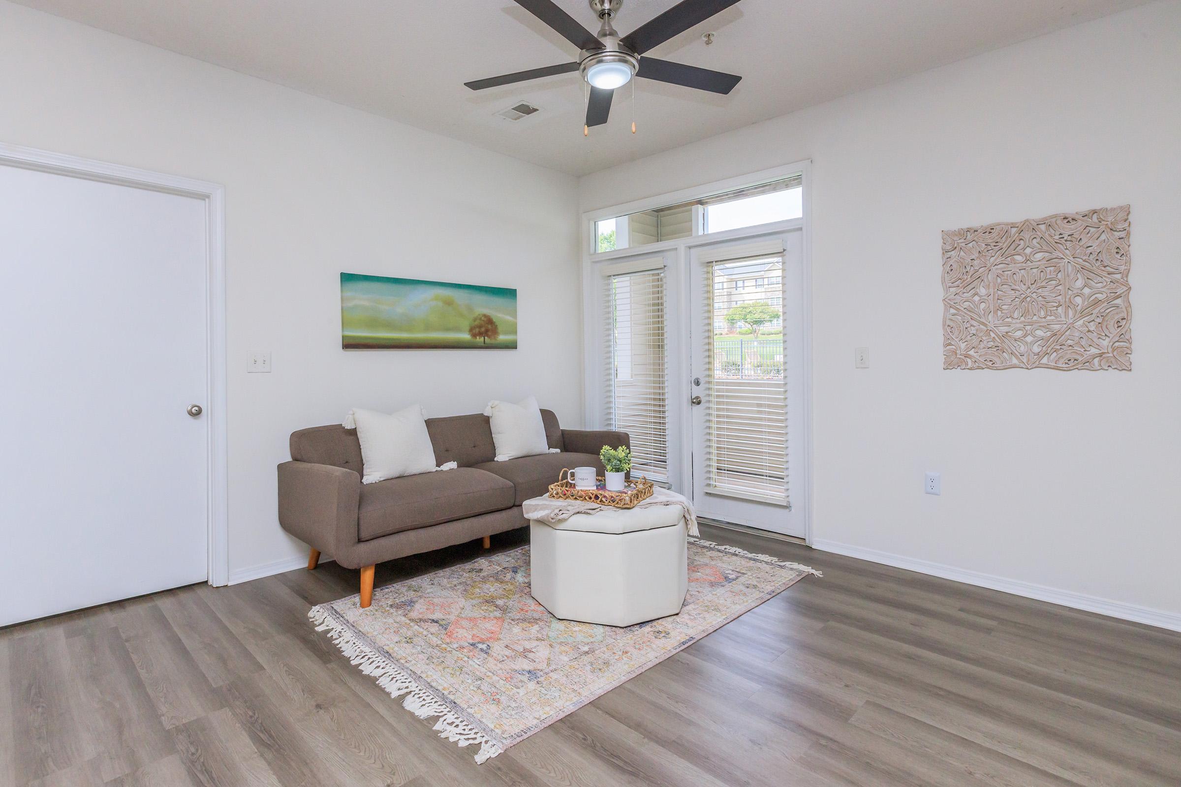 A modern living room featuring a grey sofa adorned with white cushions, a light-colored coffee table, and a colorful area rug. Natural light streams in through large windows with blinds. A wall art piece is displayed beside the door, enhancing the minimalist decor. A ceiling fan adds a stylish touch.