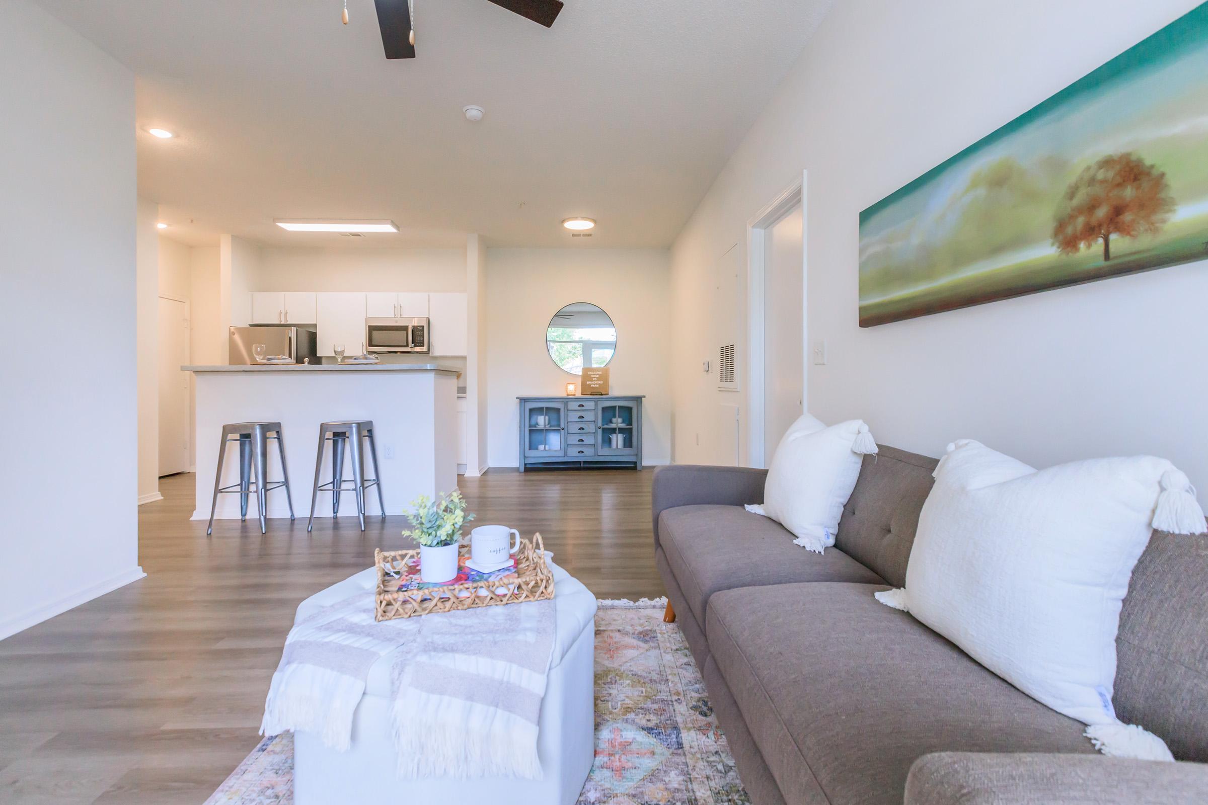 A modern living space featuring a gray couch with white pillows, a coffee table with a tray, and a colorful rug. In the background, there's a kitchen area with bar stools and appliances. A circular mirror and a wooden cabinet enhance the decor. Soft lighting and a painting of a tree complete the inviting atmosphere.