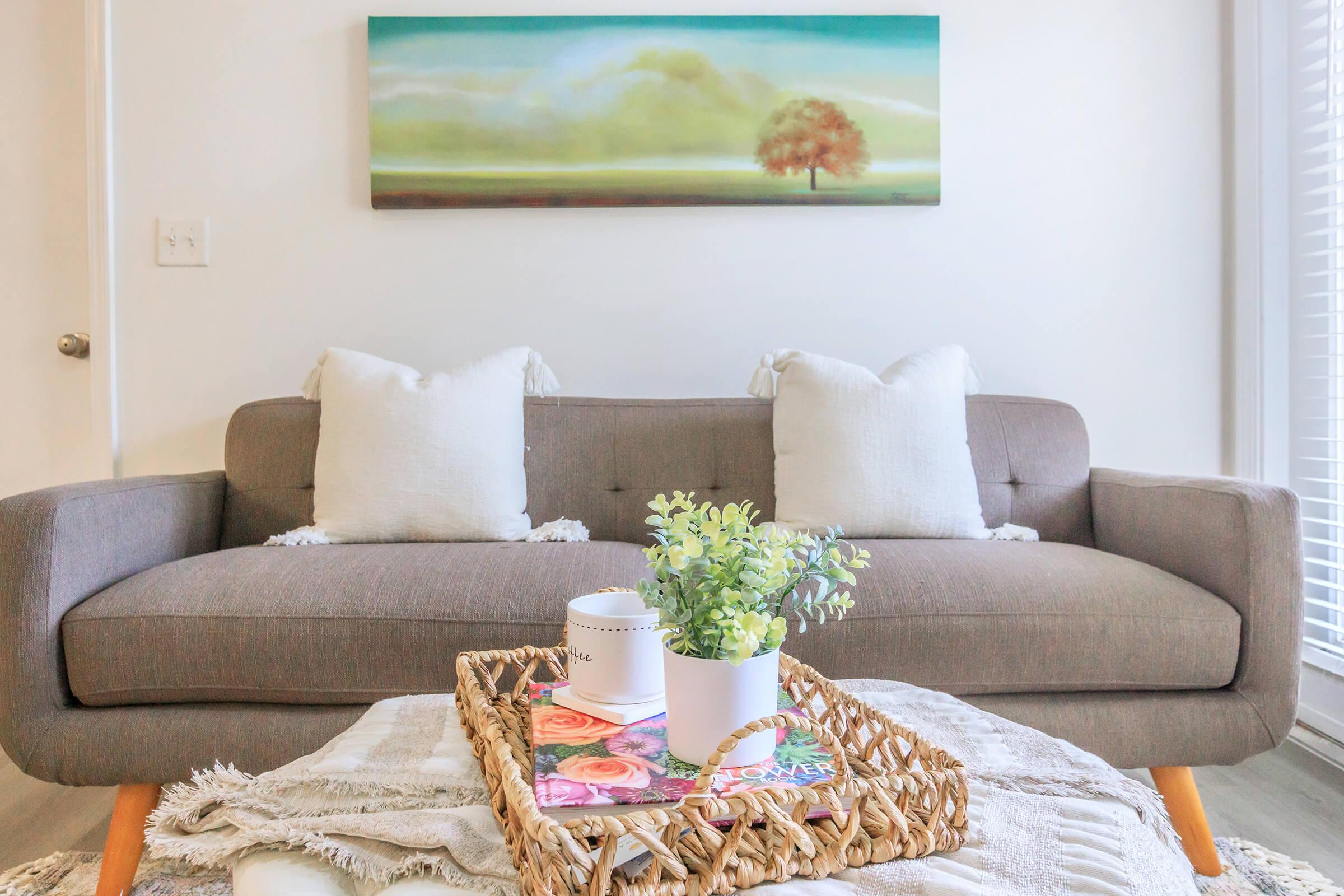 A cozy living room scene featuring a gray sofa with two white accent pillows. In front of the sofa, there is a woven tray displaying a potted plant, a decorative candle, and magazines, all set on a light-colored coffee table. A serene landscape painting hangs above the sofa, adding a touch of nature to the space.