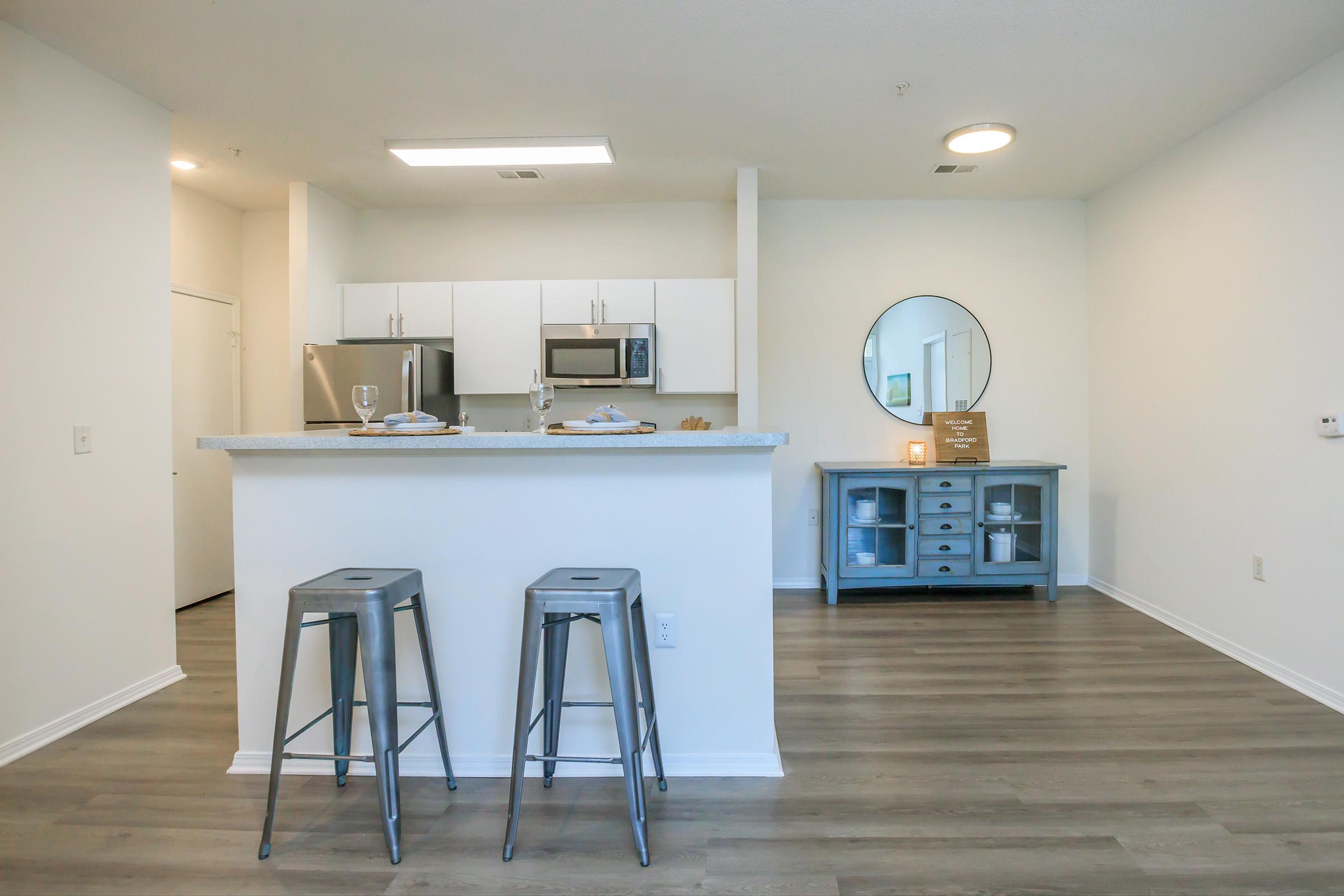 Modern kitchen with a breakfast bar featuring two metal stools and a countertop. White cabinets and a microwave are visible. In the background, a round mirror hangs on the wall above a blue-gray side table with decorative items. The flooring is light wood, creating a bright and airy atmosphere.
