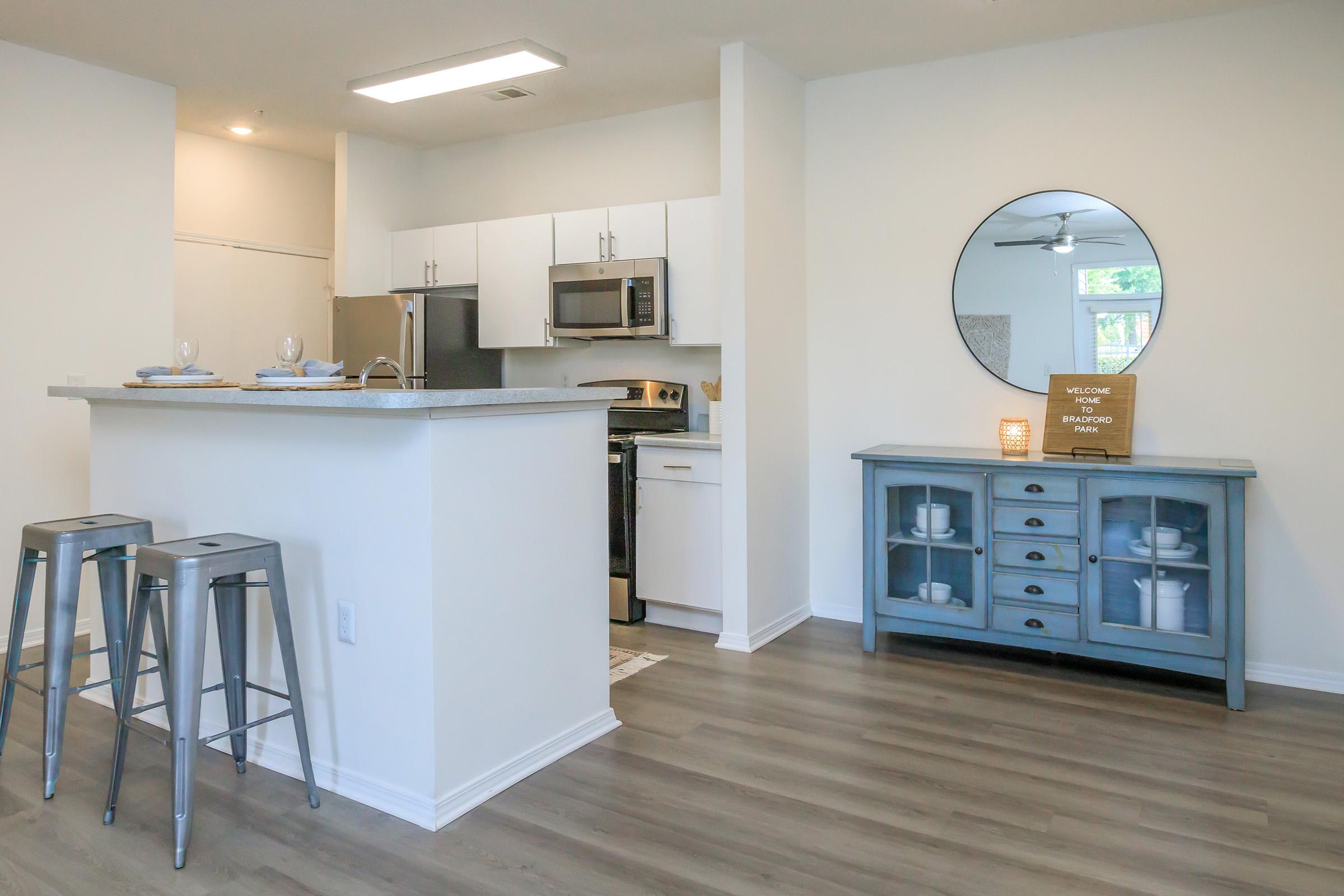 A modern kitchen and dining area featuring stainless steel appliances, a granite countertop with bar stools, and a decorative cabinet. The space is well-lit with natural light, showcasing a welcoming ambiance and a circular mirror on the wall.