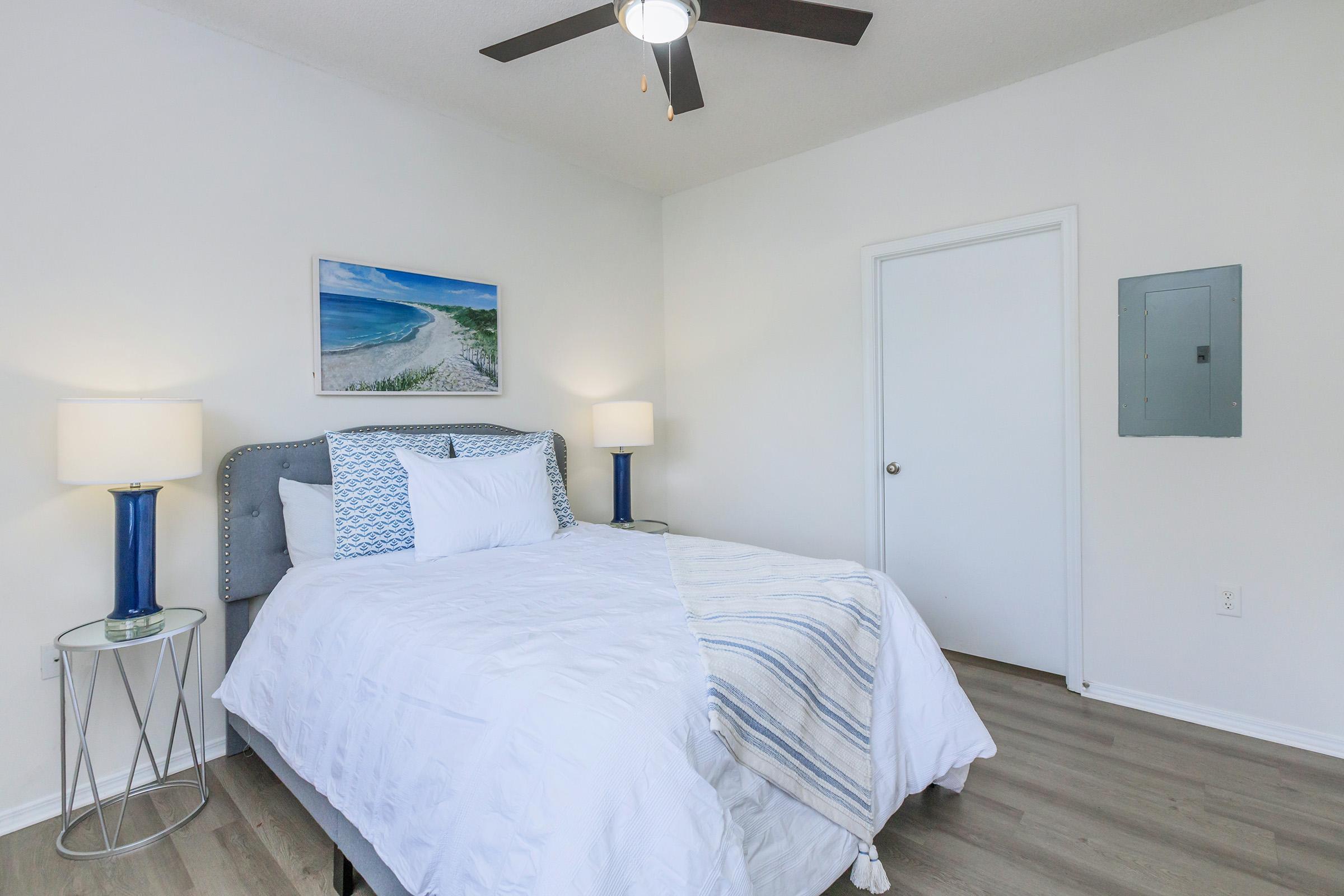 A bright and airy bedroom featuring a queen-size bed with white bedding and blue accents. Two bedside tables with blue lamps flank the bed. A beach-themed painting hangs above the headboard. The room has light wood flooring and a white door leading to another space, with a ceiling fan providing ventilation.