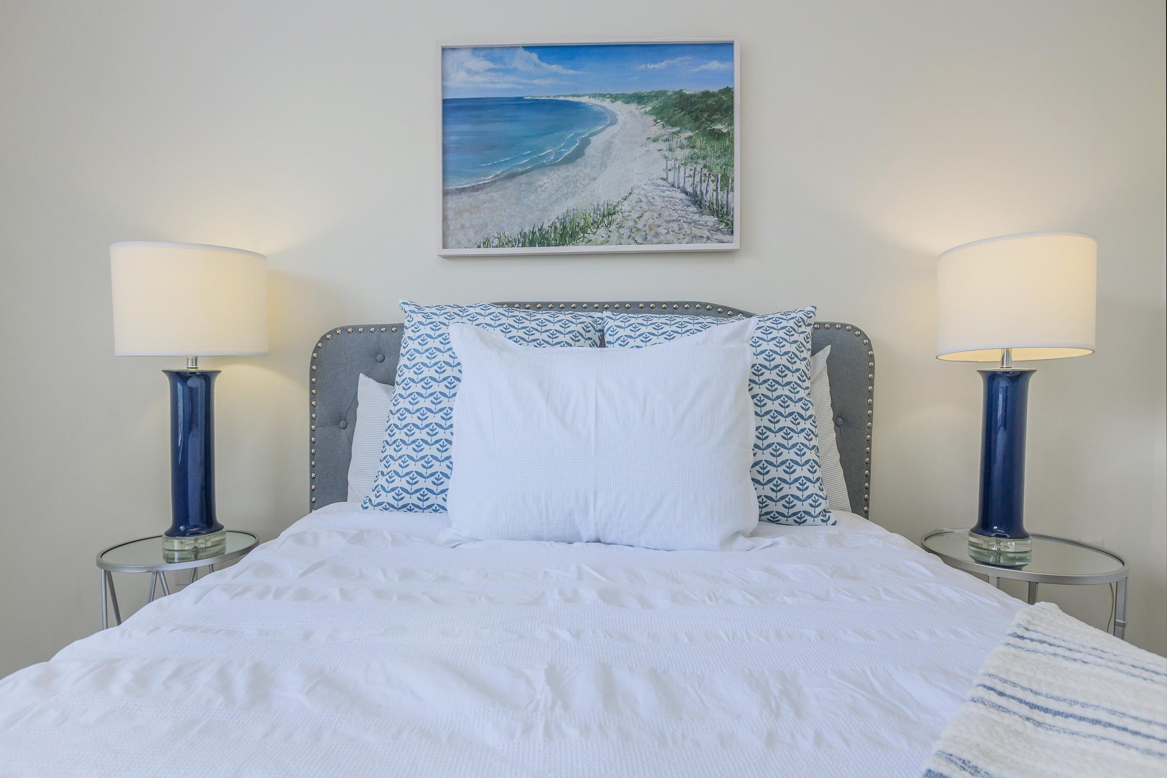 A neatly made bed with white linens and decorative blue pillows is centered in a bright bedroom. On either side of the bed, there are glass side tables topped with blue lamps. Above the bed, a painting depicts a tranquil beach scene, adding a serene touch to the room's decor.