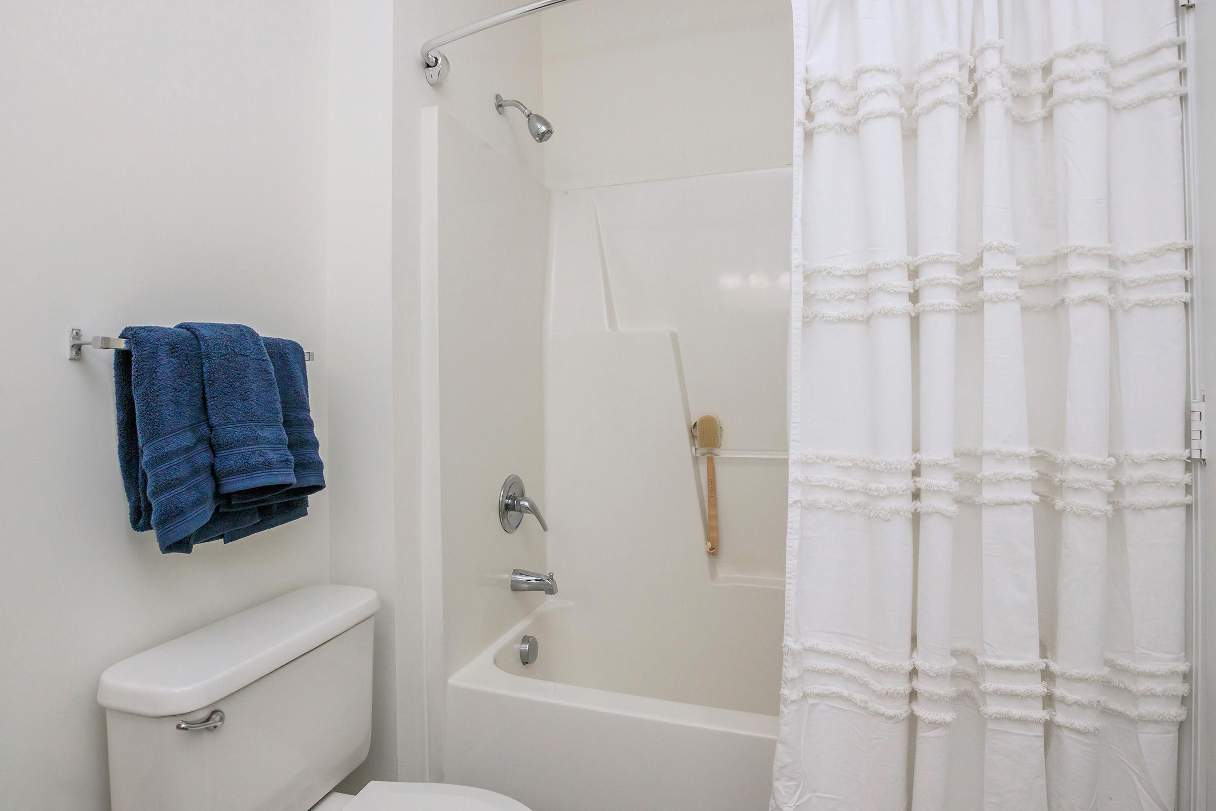 A clean bathroom scene featuring a bathtub with a shower, a white shower curtain, and neatly folded blue towels hanging on a towel rack. The toilet is visible beside the bathtub, contributing to a tidy and organized space.