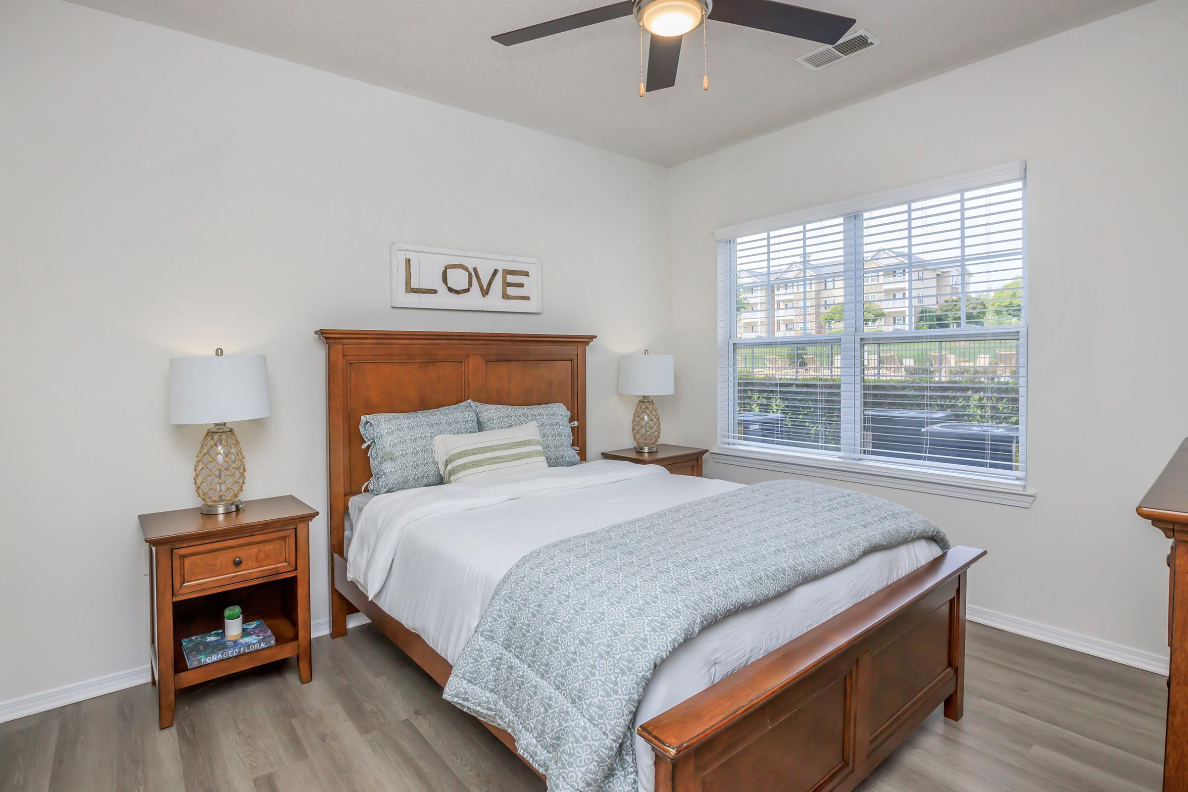 A cozy bedroom featuring a wooden bed with white bedding and decorative pillows. There are two bedside tables with lamps, a decorative sign that says "LOVE" on the wall, and a window with blinds allowing natural light in. The room has light-colored walls and a warm wooden floor.
