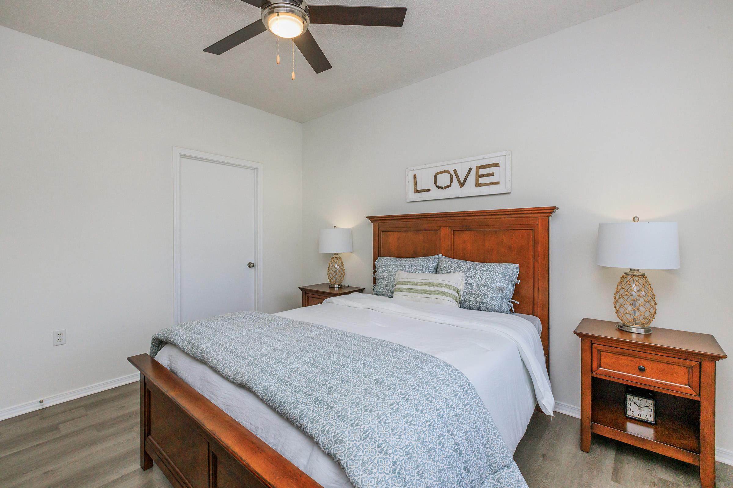 Cozy bedroom featuring a queen-sized bed with white bedding and a decorative blue throw. Two bedside tables with decorative lamps flank the bed. A wall sign that reads "LOVE" hangs above the headboard. The room has a light, neutral color scheme and a modern ceiling fan.