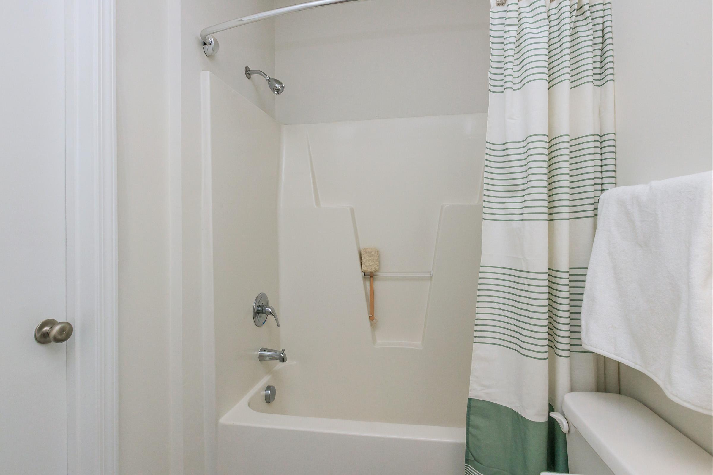 A clean shower area featuring a white tub and wall, a stainless steel showerhead, and a light-colored striped shower curtain. A bamboo bath brush hangs inside the tub, and a white towel is neatly placed on a nearby shelf. The space is bright and well-maintained, emphasizing a minimalist design.