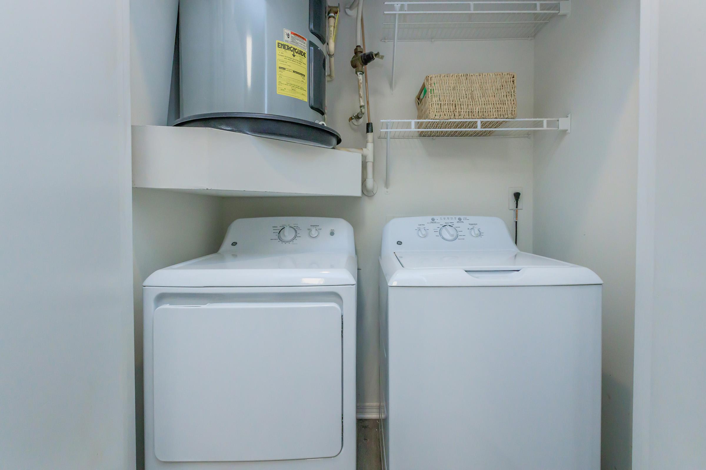 A laundry room featuring a white washing machine and a white dryer side by side. Above them is a water heater with a warning label. There is a metal shelf above with a wicker basket for storage, and the walls are painted in a light color, creating a clean and organized space.