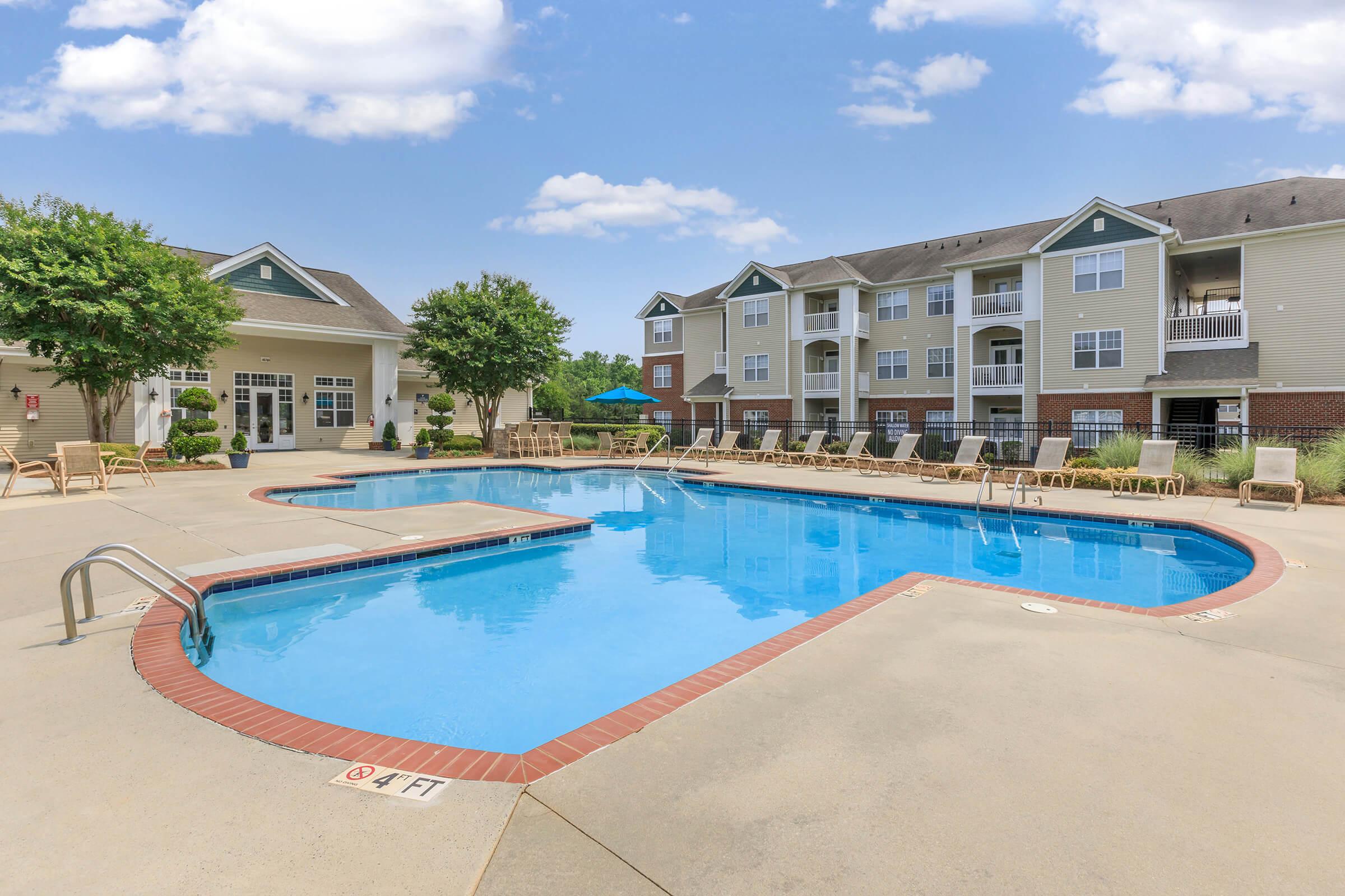 A clear blue swimming pool surrounded by lounge chairs and umbrellas, with a backdrop of modern apartment buildings. Lush greenery and trees line the area, offering a relaxing outdoor space. The sky is bright with a few white clouds, enhancing the inviting atmosphere of the poolside area.