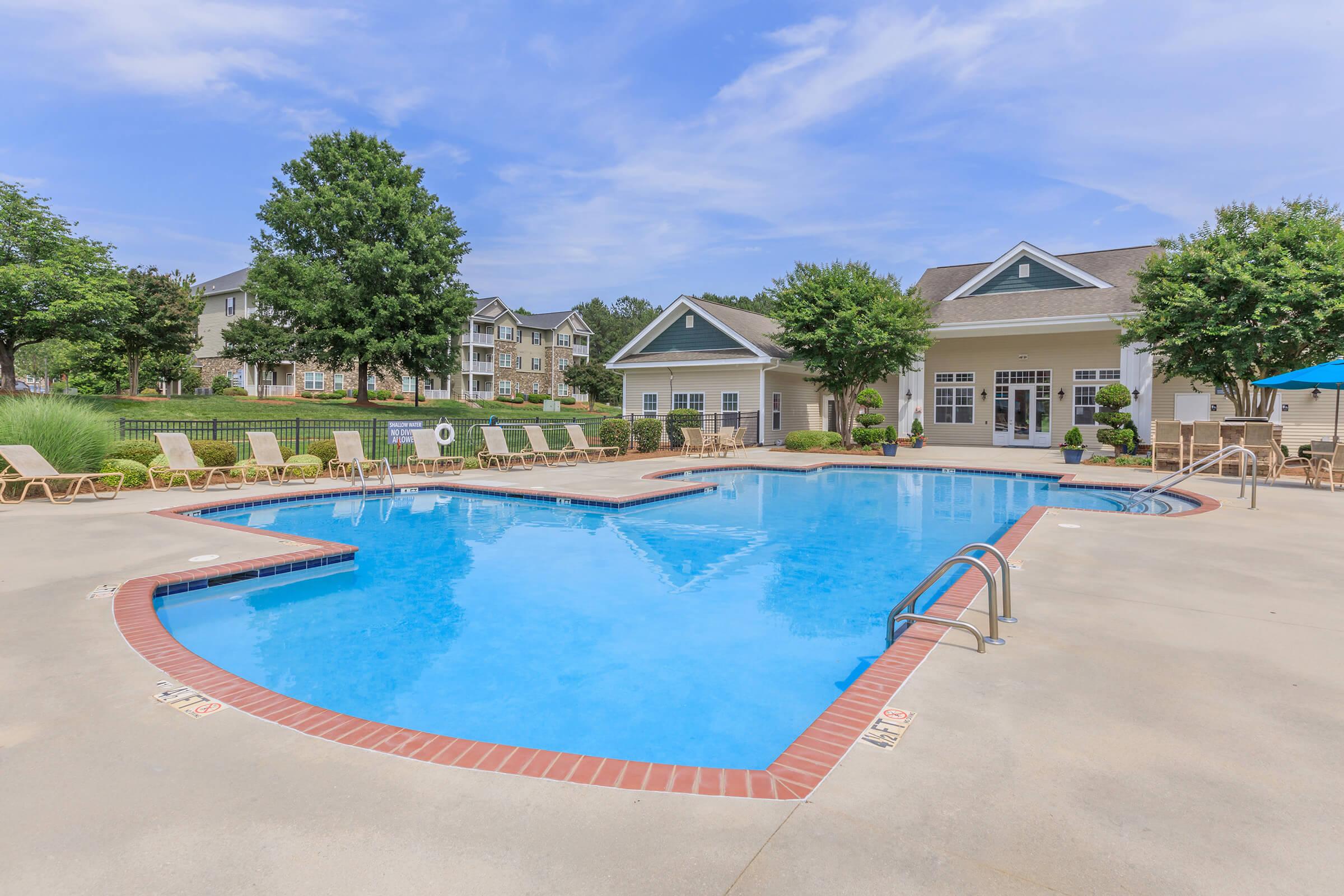 A sparkling blue swimming pool surrounded by lounge chairs, with lush greenery and trees in the background. The pool area features a gently curved shape and is accompanied by a building with large windows, creating a relaxing outdoor space in a residential setting.