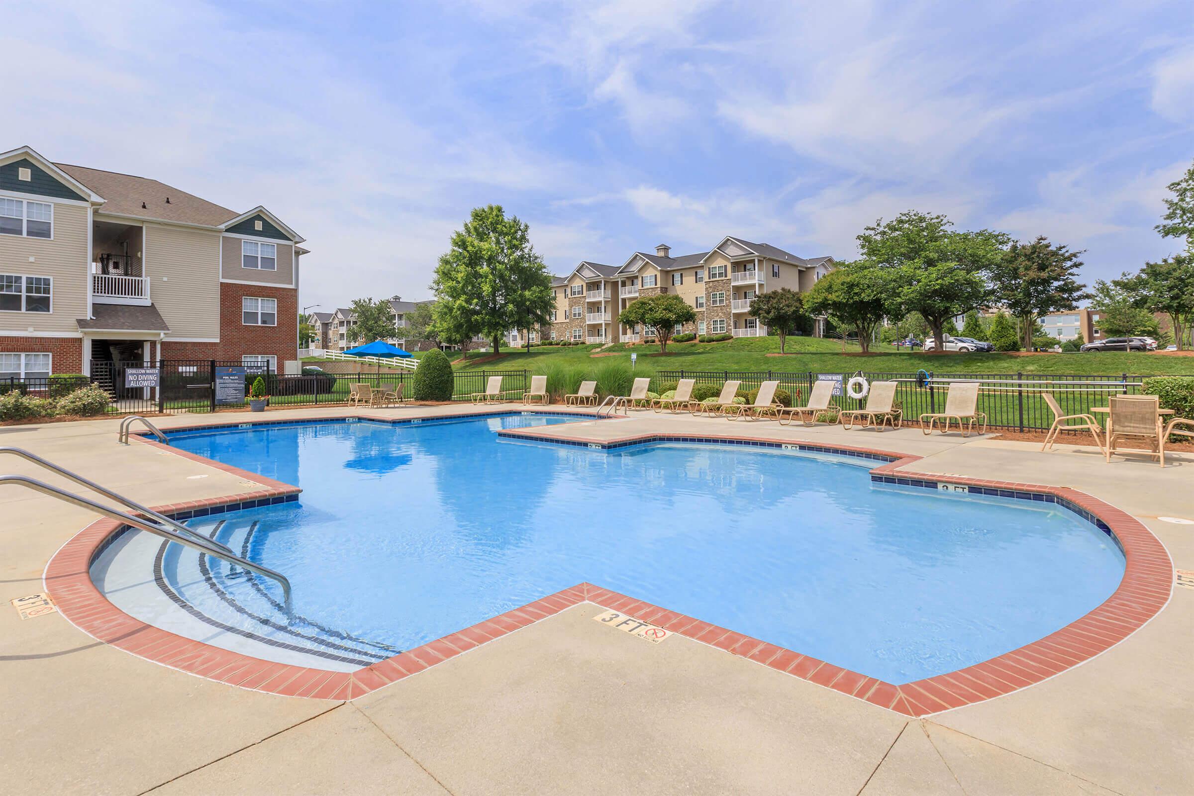A serene swimming pool surrounded by lounge chairs, with green lawns and residential buildings in the background. The sky is partly cloudy, adding to the peaceful atmosphere. Perfect for relaxation and leisure activities.