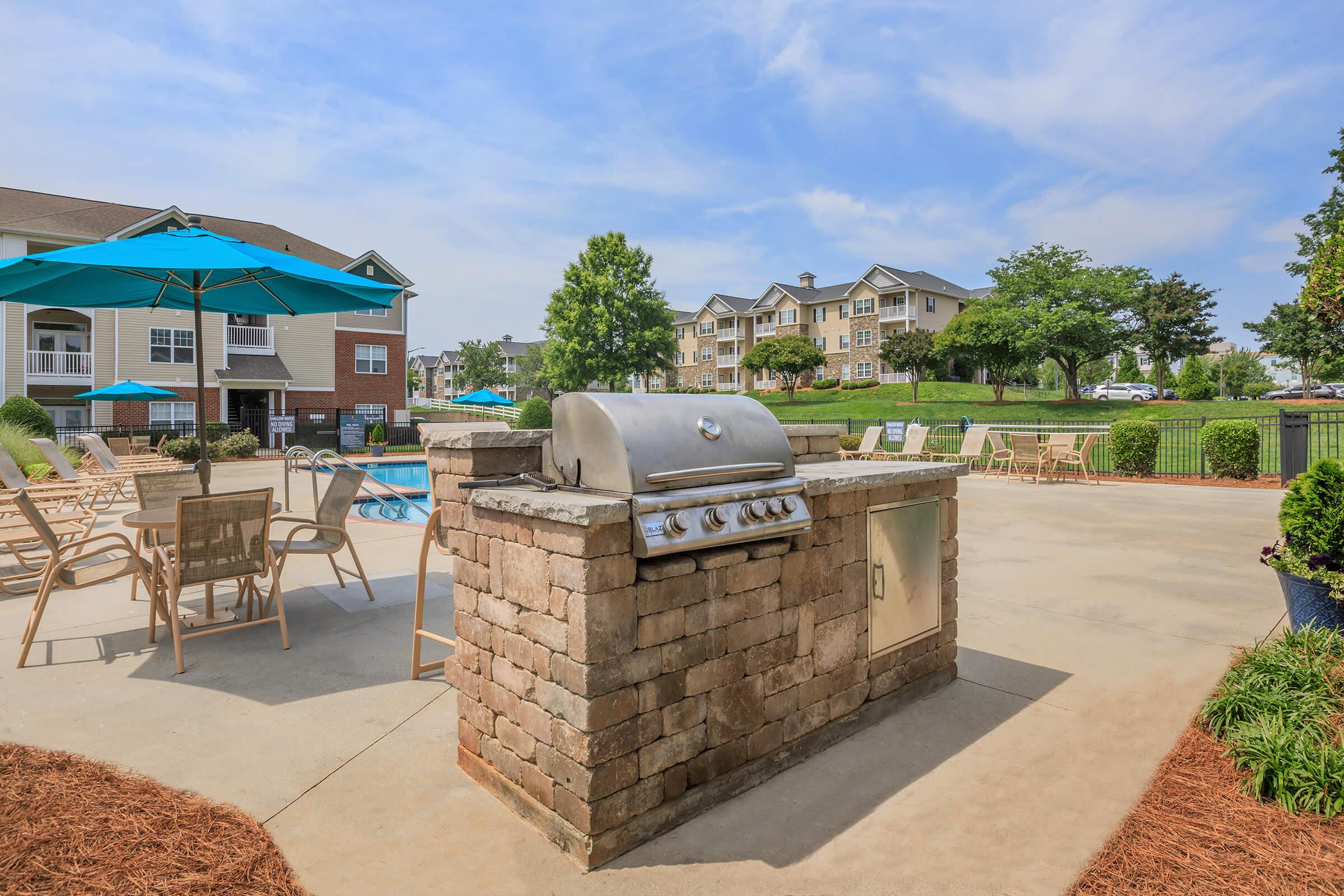 Grill station made of stone in a poolside setting, surrounded by lounge chairs and umbrellas. In the background, there are residential buildings and green trees under a clear blue sky. The pool area is ready for recreational use and relaxation.