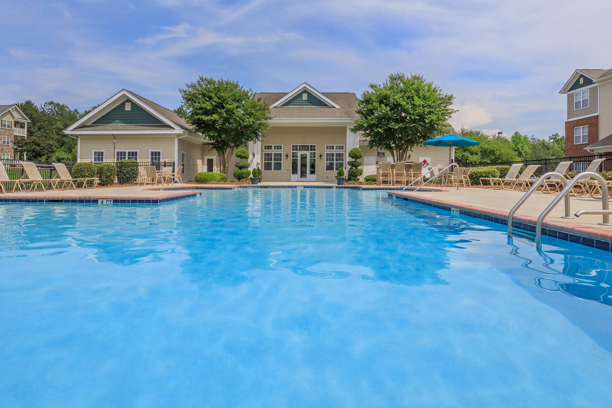 A clear blue swimming pool surrounded by lounge chairs and a well-maintained lawn. In the background, a clubhouse with large windows and a covered patio area is visible. The scene is bright and inviting, with trees and landscaping creating a relaxing atmosphere.